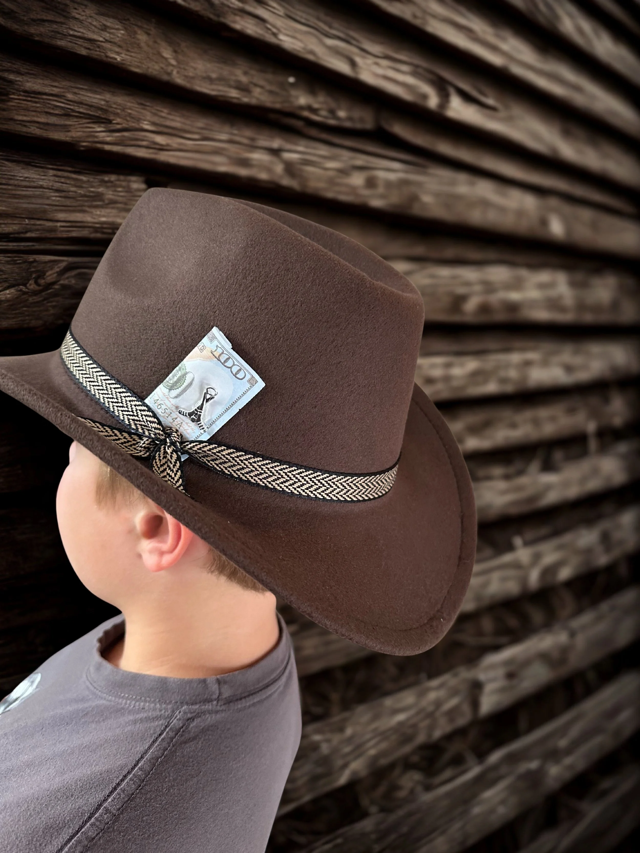A child wearing a brown cowboy hat with a dollar bill tucked into the hatband, standing against a wooden wall.