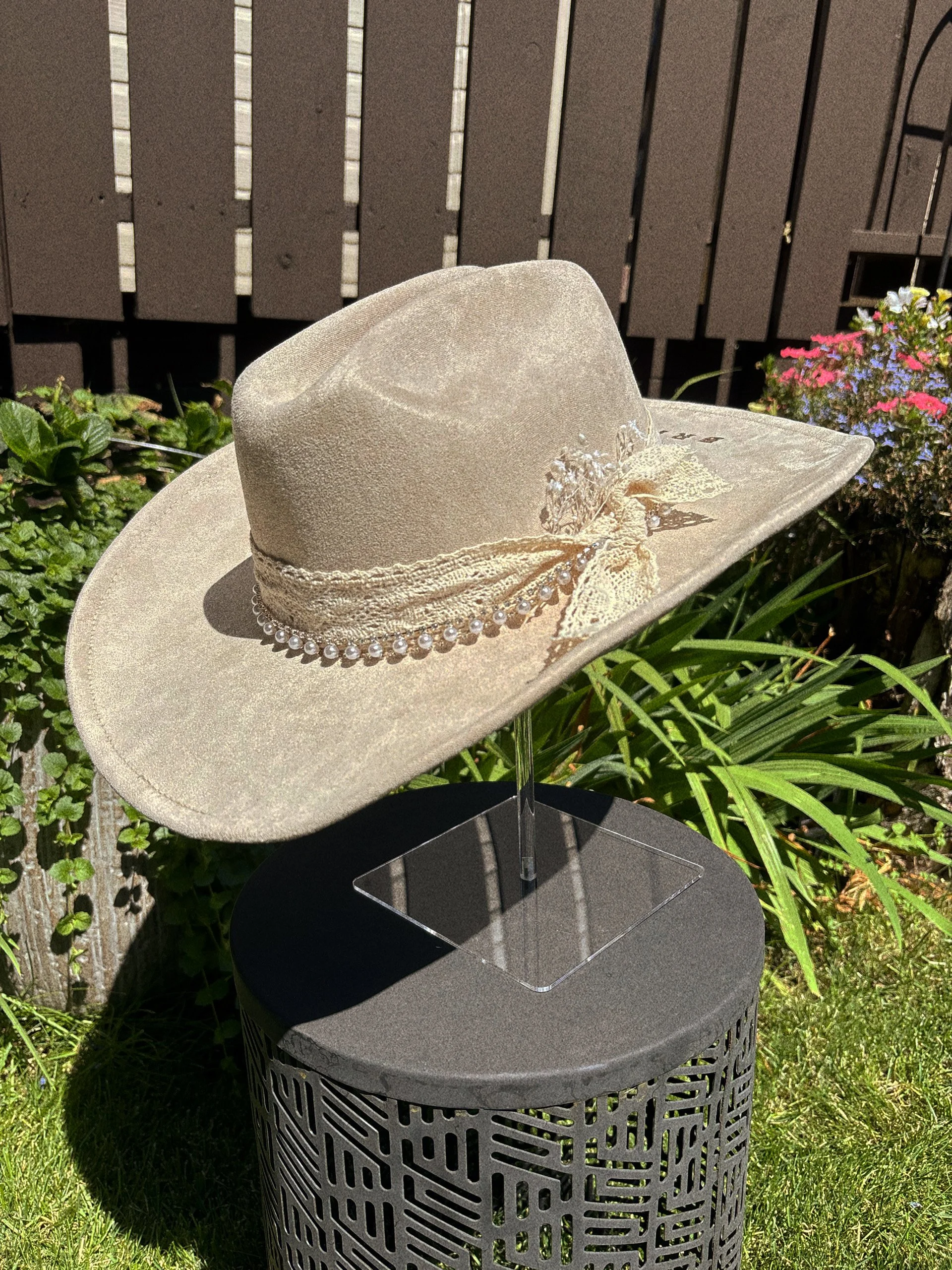 A beige cowboy hat with a beaded band and lace decoration displayed outdoors on a black pedestal, with green plants and a wooden fence in the background.