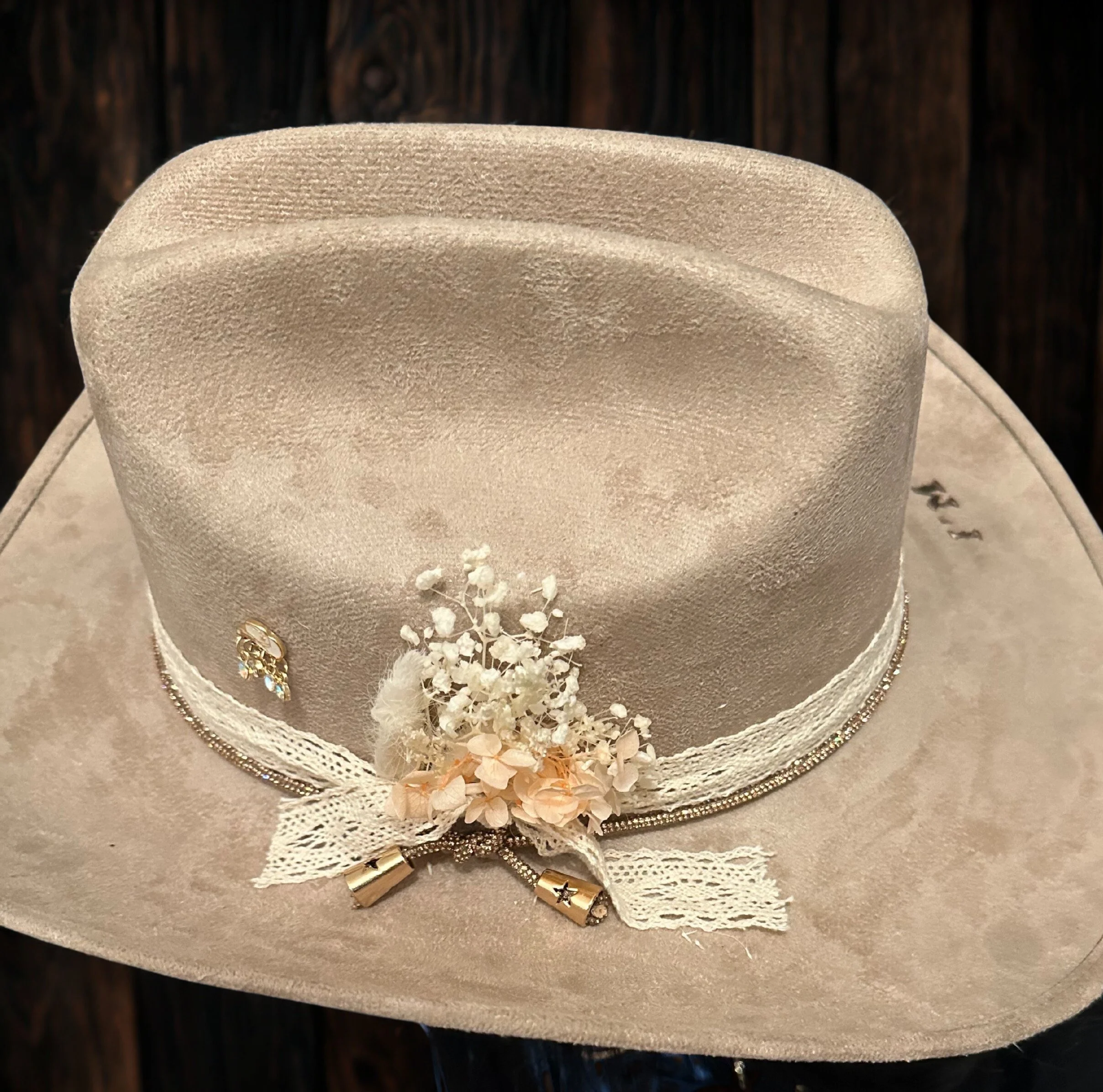A cream-colored felt cowboy hat decorated with lace ribbon, small white flowers, and metallic charms, displayed on a light-colored surface with a dark wooden background.