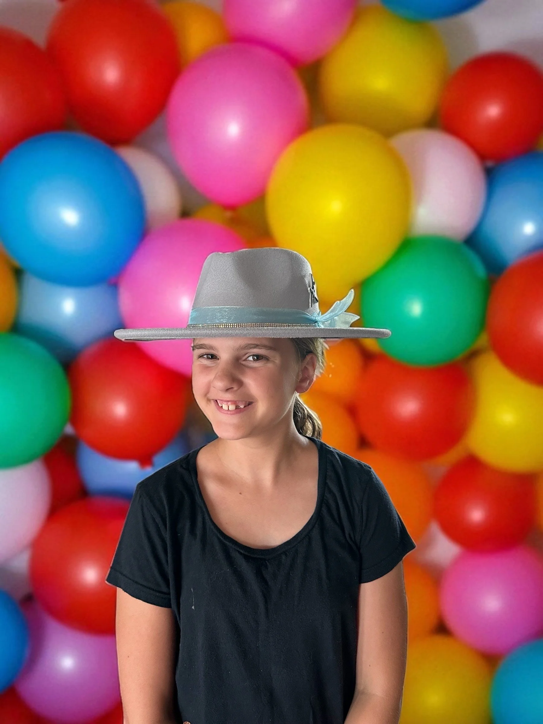 A smiling girl wearing a gray fedora hat with a blue ribbon, standing in front of a colorful background of pink, yellow, blue, green, orange, and red balloons.