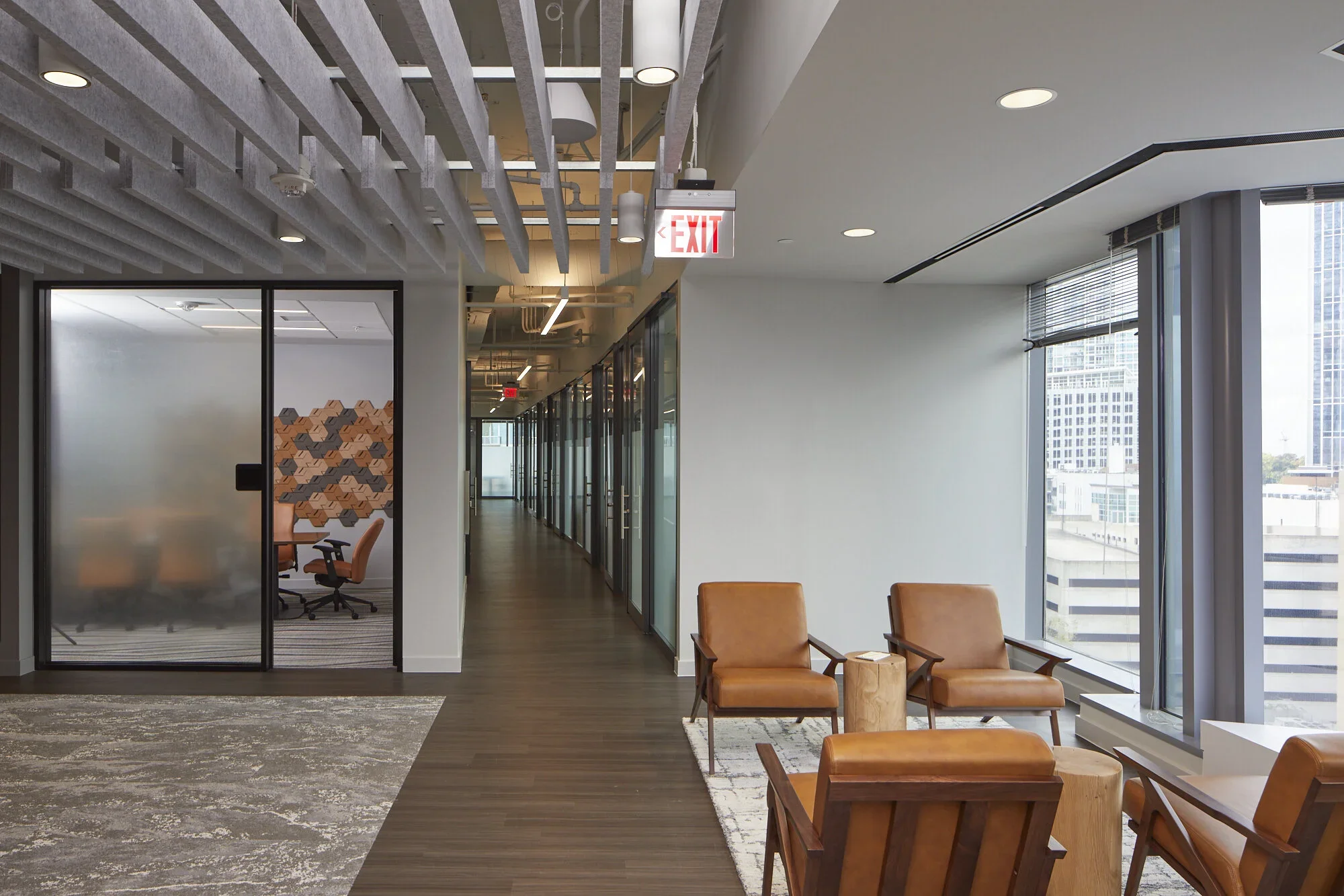 Modern office lounge area with brown chairs, a wooden side table, large windows showing city buildings, a carpeted living space, and glass-walled conference room.