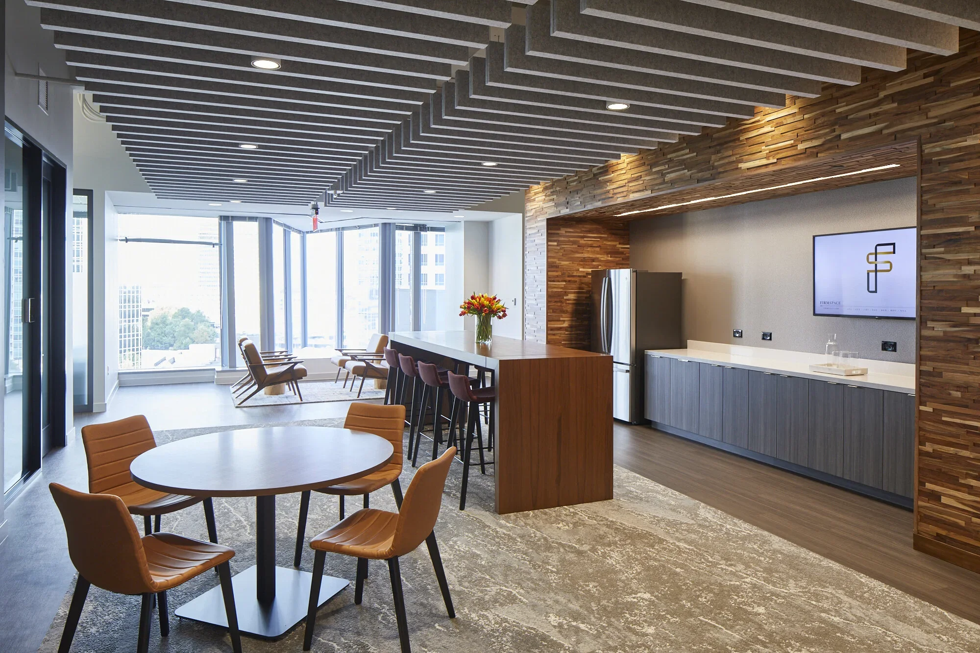 Modern lounge area with wooden ceiling and wall accents, a round table with six tan chairs, a bar with five barstools, and a kitchenette with a refrigerator, countertop, and a wall-mounted TV, large windows in the background letting in natural light.