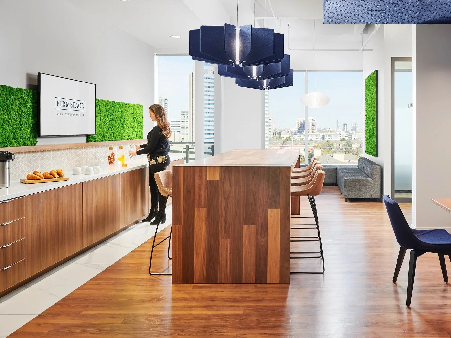Modern office break room with a woman preparing coffee, wooden counter, large window with city view, green wall decor, and seating area.