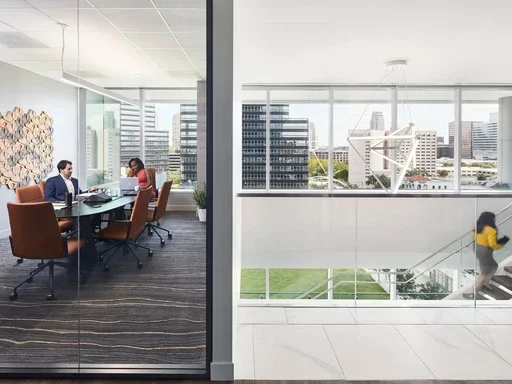 Office meeting room with three people sitting at a conference table and a woman walking downstairs in a modern office building with city view.