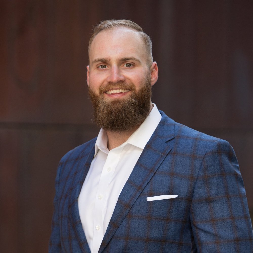 Portrait of a smiling man with a beard, wearing a blue plaid suit and white shirt, standing against a dark background.