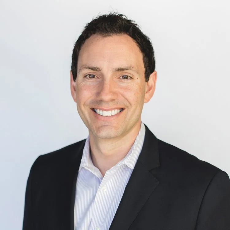 Professional headshot of a smiling man with dark hair, wearing a black suit and a white collared shirt, against a plain white background.