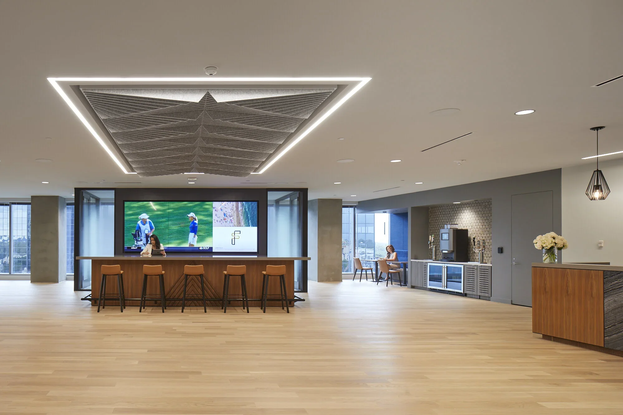 Modern hotel lobby with a bar area, a large digital screen, tables, and chairs, featuring wooden flooring and minimalist decor.