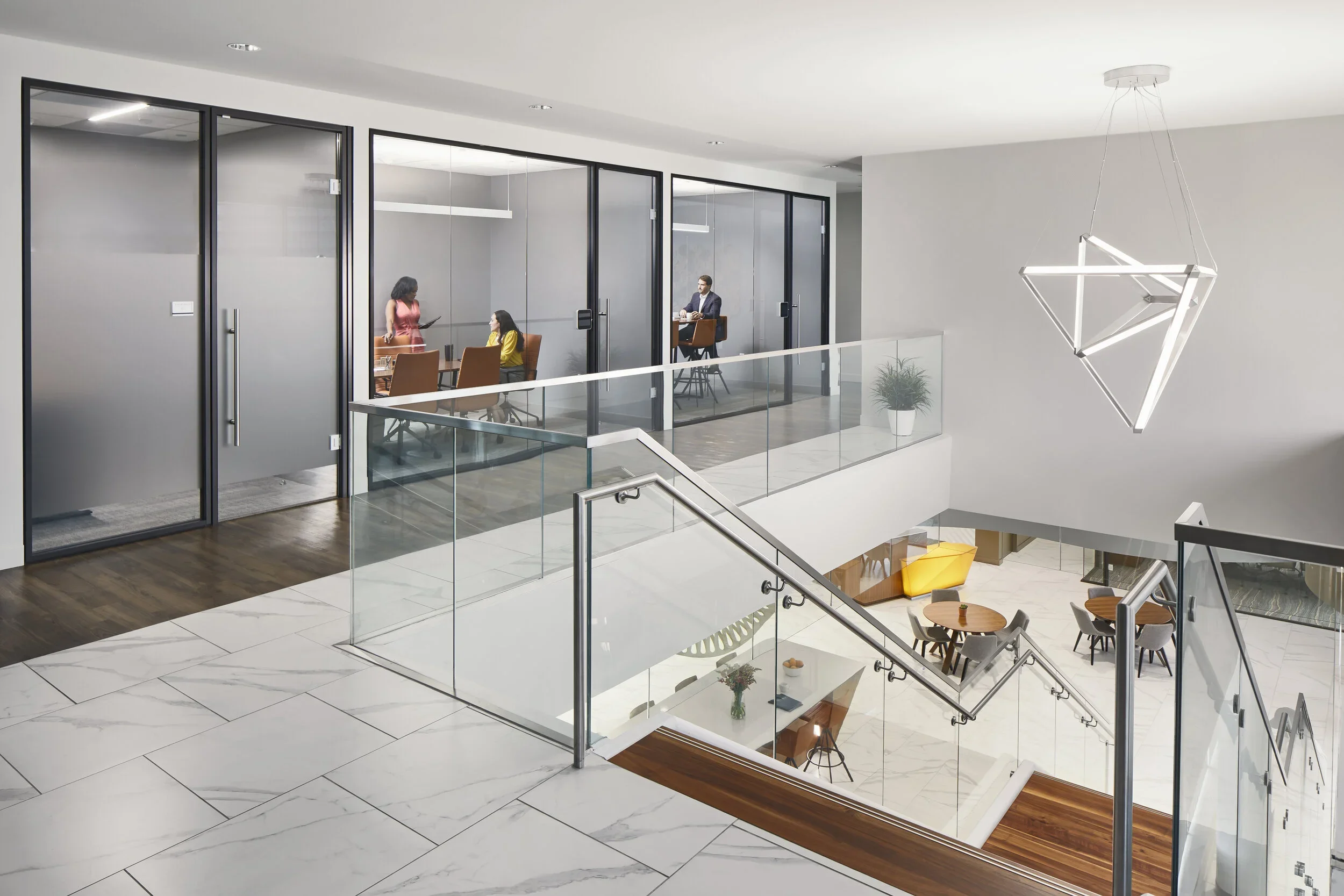 Modern office interior with glass walls, a staircase, and seating areas, including round tables with chairs and a pendant light fixture.