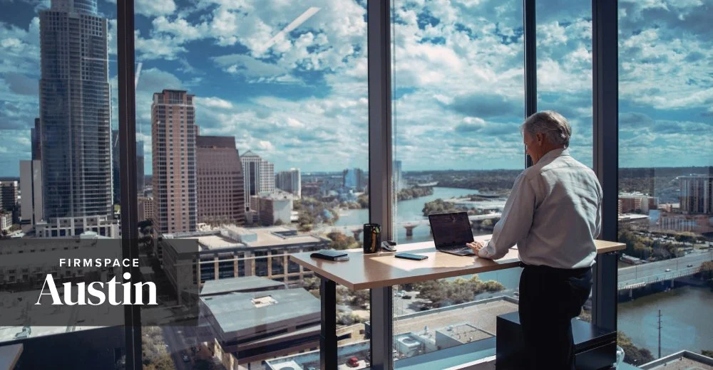 An older man in a white shirt working on a laptop at a high-rise office with large windows, overlooking a city skyline and river in Austin, Texas.