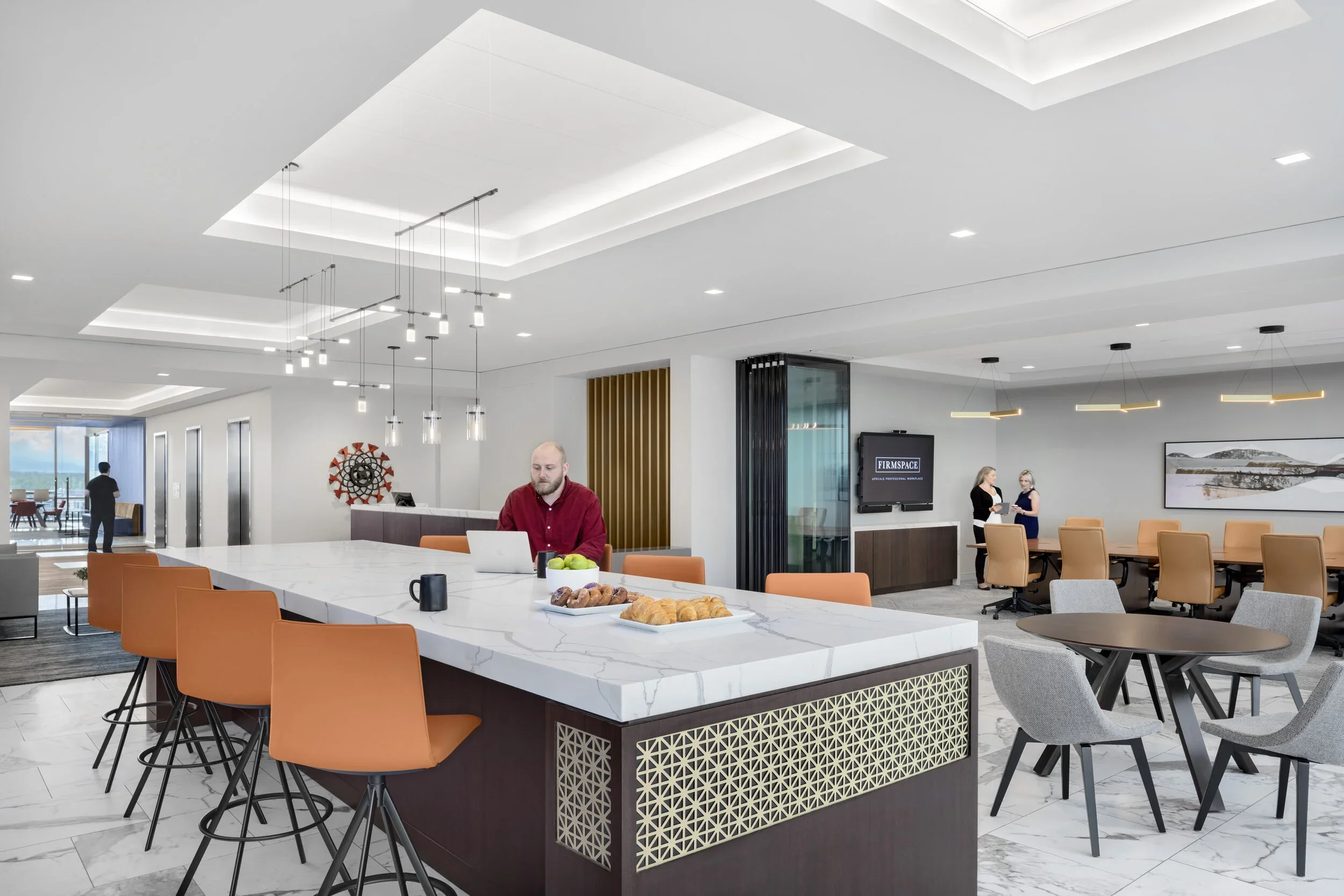 Modern office lounge area with a large central marble countertop, orange bar stools, a man working on a laptop, and a conference space in the background with two women talking near a wall-mounted TV.