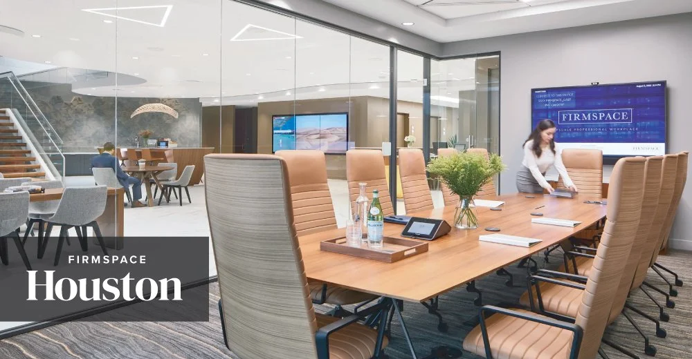 A modern conference room in a corporate office with a long wooden table, beige office chairs, and a woman cleaning the table. There is a large screen on the wall displaying 'FIRMSPACE Houston' and glass walls showing a seating area with additional chairs and tables.