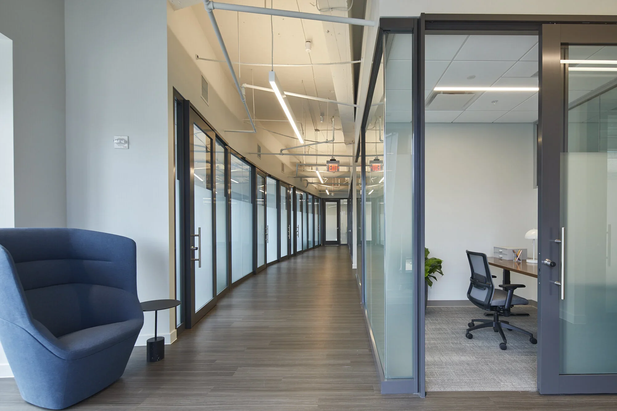 Modern office corridor with glass-walled meeting rooms on the right, blue lounge chair and small table on the left, and wooden flooring and ceiling with exposed pipes and lighting.