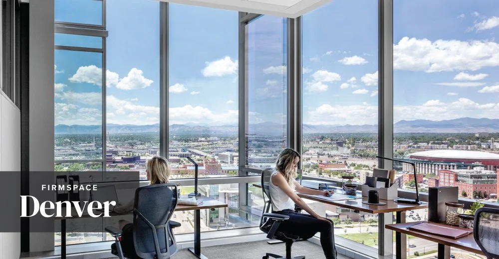 Modern office space with large glass windows overlooking Denver cityscape and mountains, with two women working at desks.