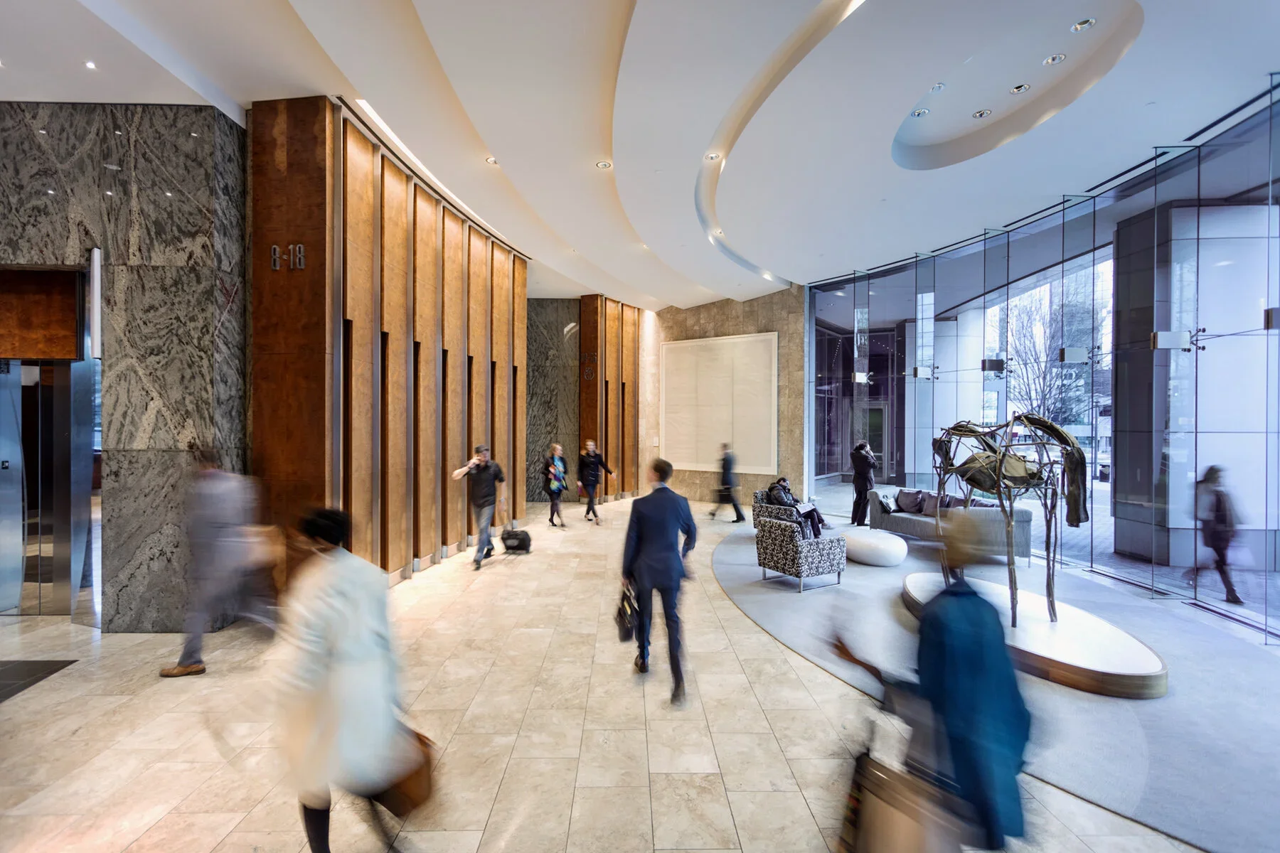 Lobby of a hotel with people walking and sitting, modern interior with marble walls, wooden panels, large glass windows, and contemporary furniture.