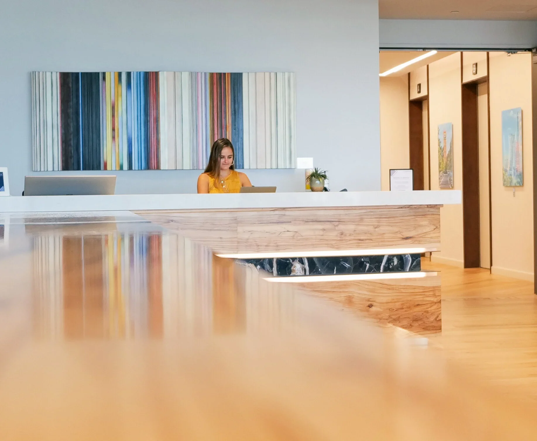 A hotel or office reception area with a woman standing behind a white reception desk, engaging with her tablet, and framed abstract art hanging on the wall behind her.