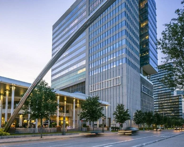 Tall modern glass office building with a supporting steel beam structure and tree-lined sidewalk in front.