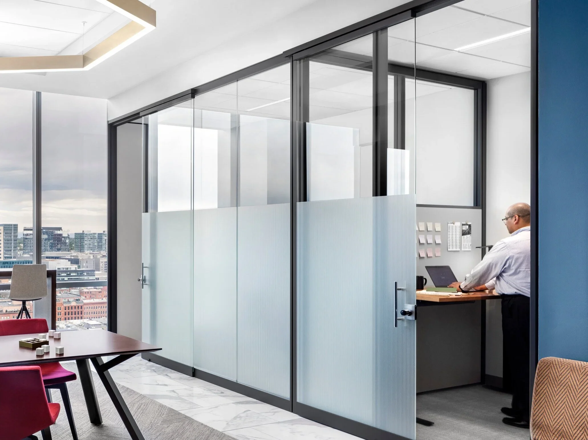Modern office space with glass-walled meeting room, a man working on a laptop, and conference table with red chairs.