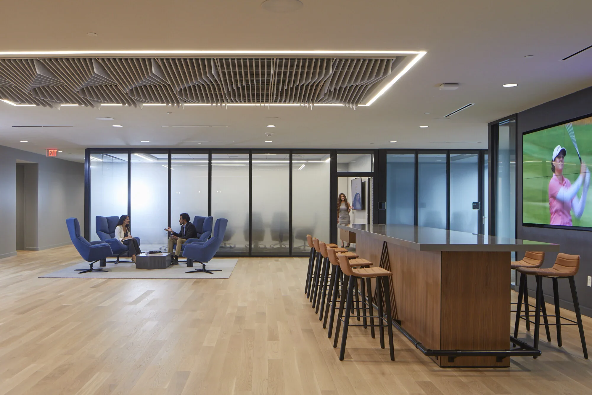 Modern office lounge with two women having a conversation, bar area with high chairs, large wall-mounted TV displaying a person playing golf, glass-walled conference room in the background.