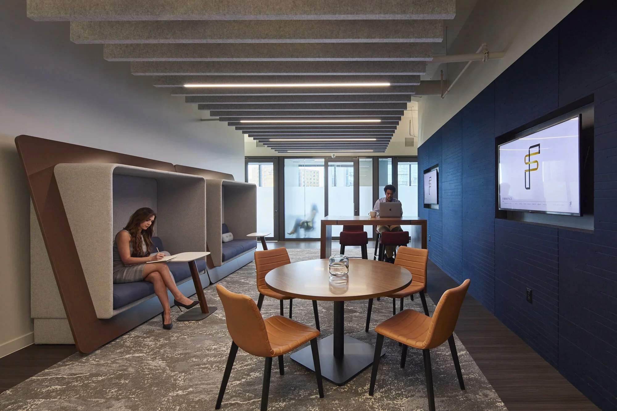 Modern office lounge area with built-in booth seating, a round table with four chairs, and a man working on a laptop at a high table. There are large windows, wall-mounted screens, and a textured ceiling.