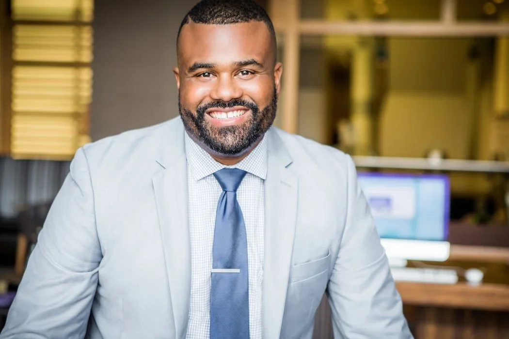 Man smiling, dressed in a light gray suit and blue tie, in office setting with computer monitors in background.