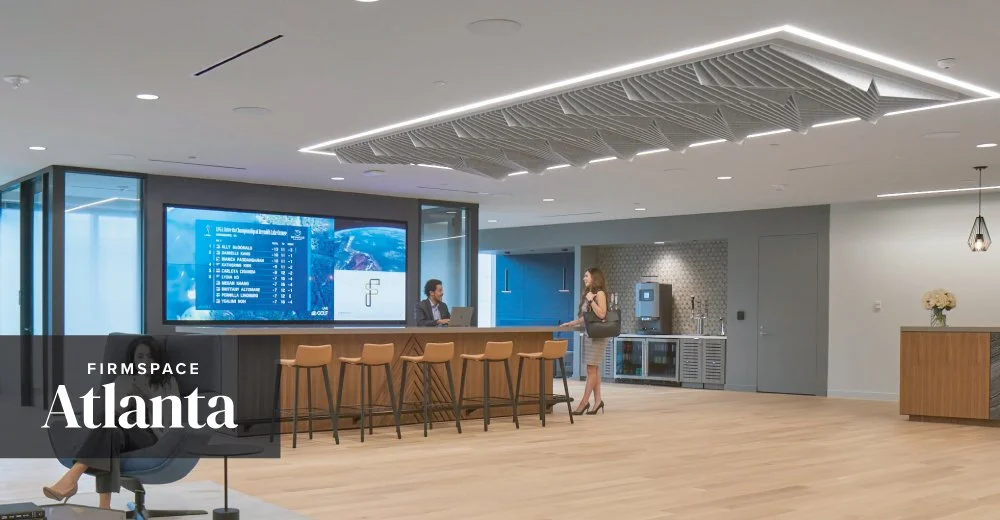 Modern office lobby with a reception desk, a large digital screen, and a woman talking to a receptionist. There is a seating area and a refreshment station in the background.