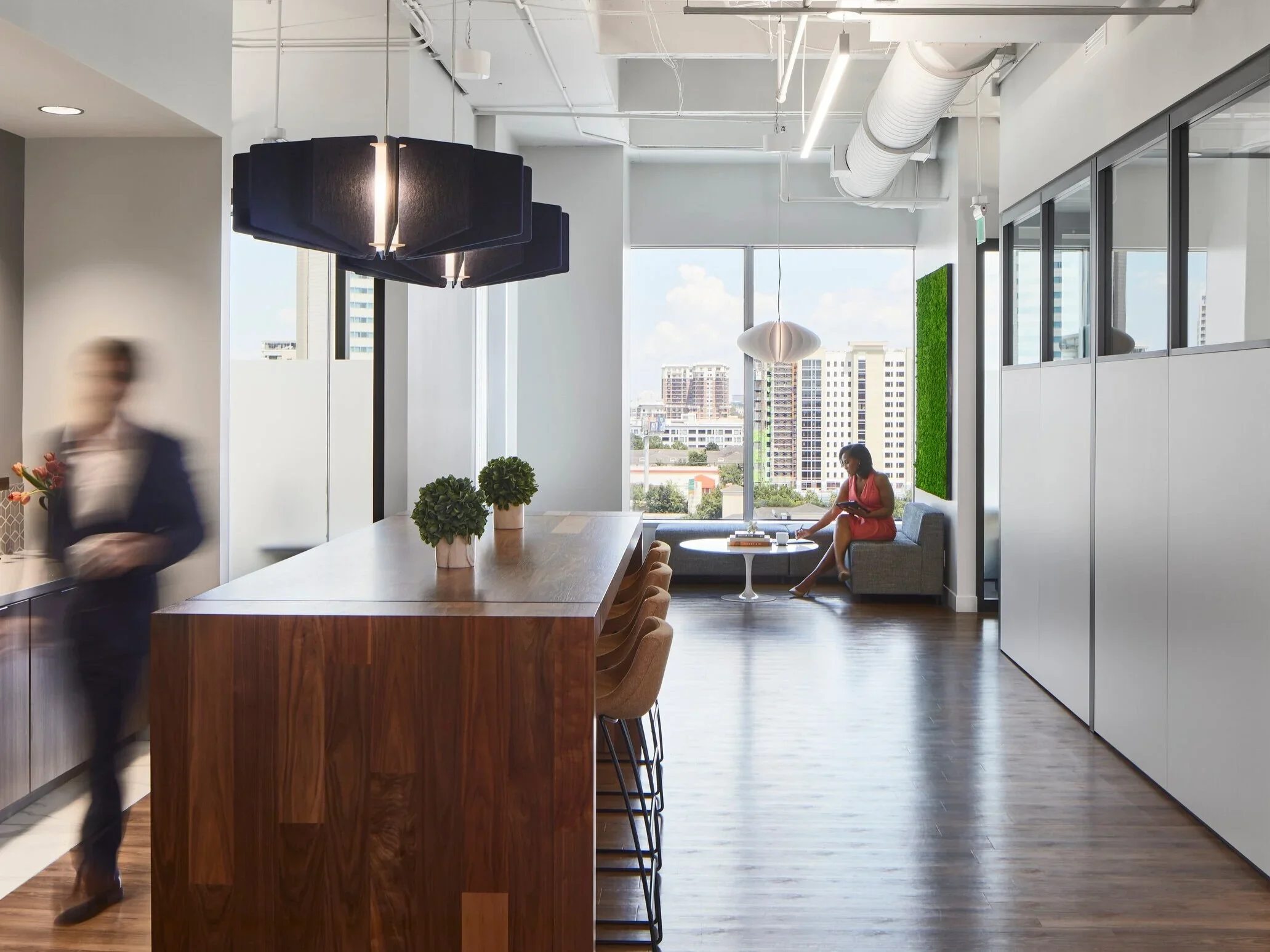 Modern office space with a large wooden counter, woman sitting on a gray couch near a large window, and a woman blurred in motion walking past. Cityscape view outside the window.