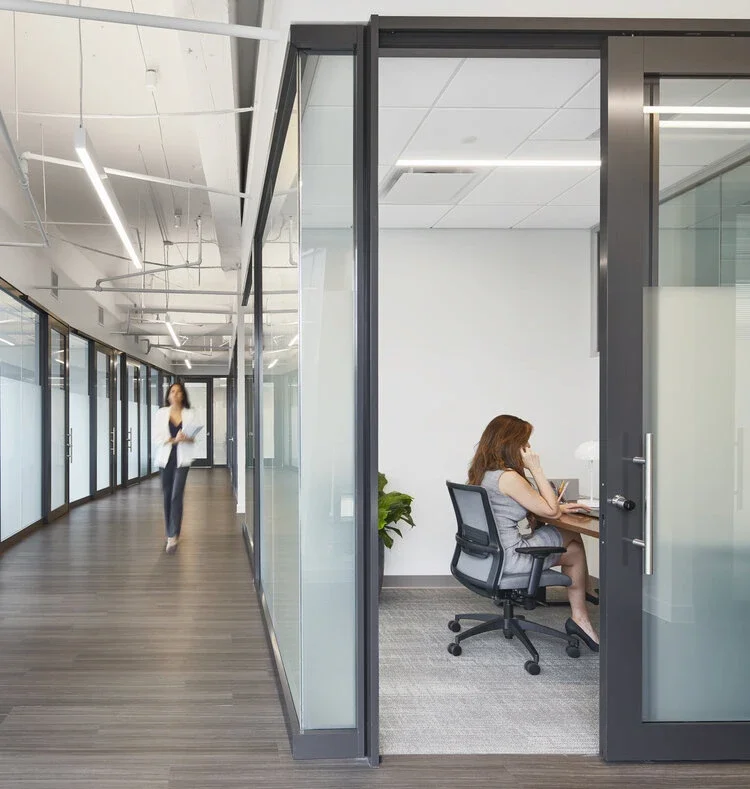 An office hallway with glass-walled offices. One woman is walking down the hallway, and another woman is sitting at a desk inside a glass office, talking on the phone or listening to a conversation.
