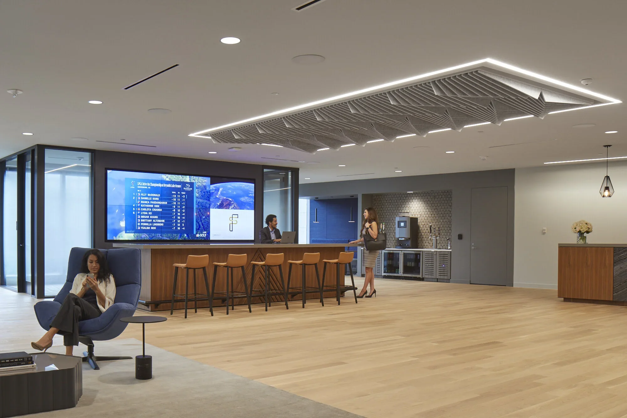 Modern hotel lobby with a bar, a large digital screen displaying golf tournament scores, a woman with a handbag talking to a bartender, a woman sitting in a blue armchair using her phone, and a water dispenser area in the background.