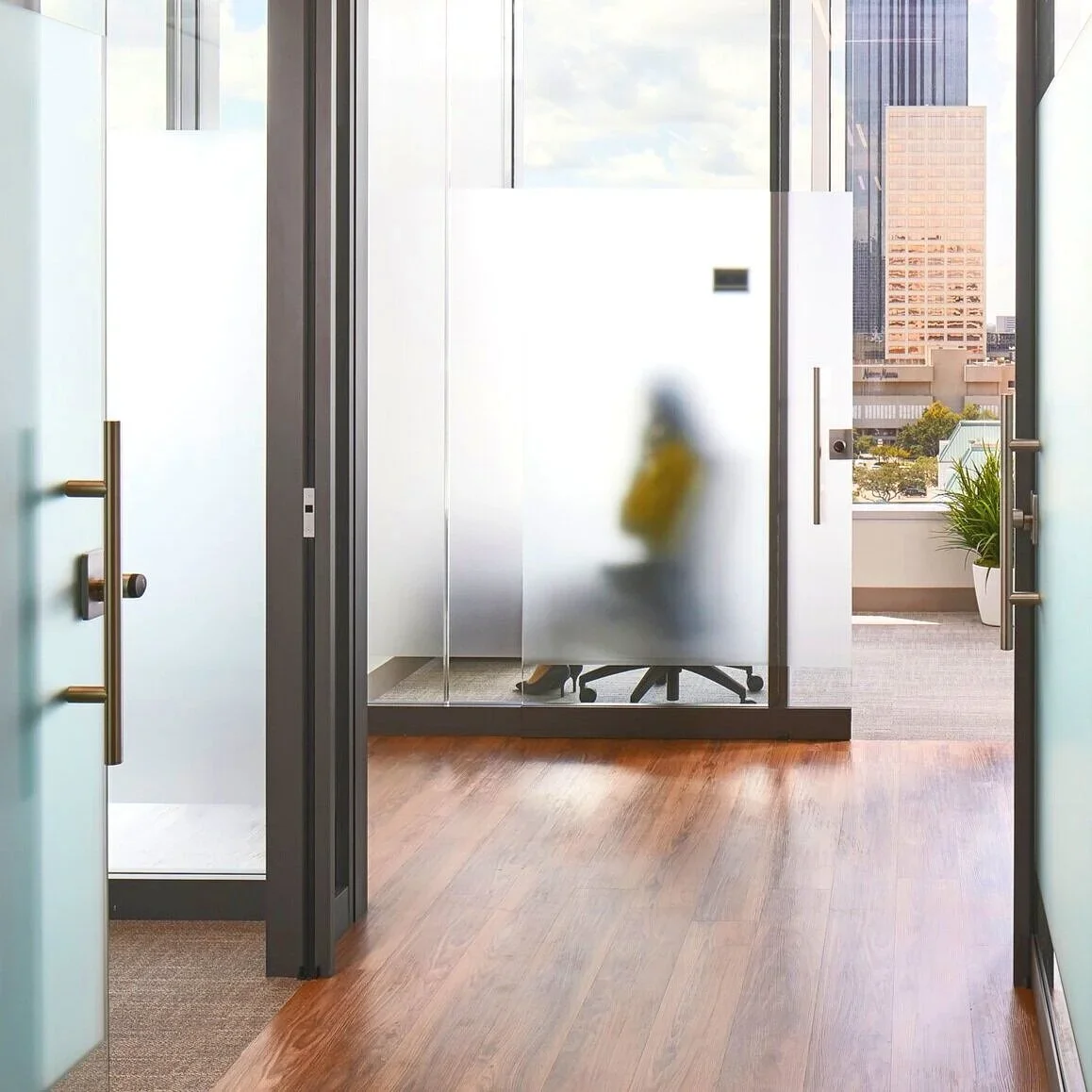 View through glass doors into a modern office with a blurred silhouette of a person sitting at a desk and city skyline in the background.
