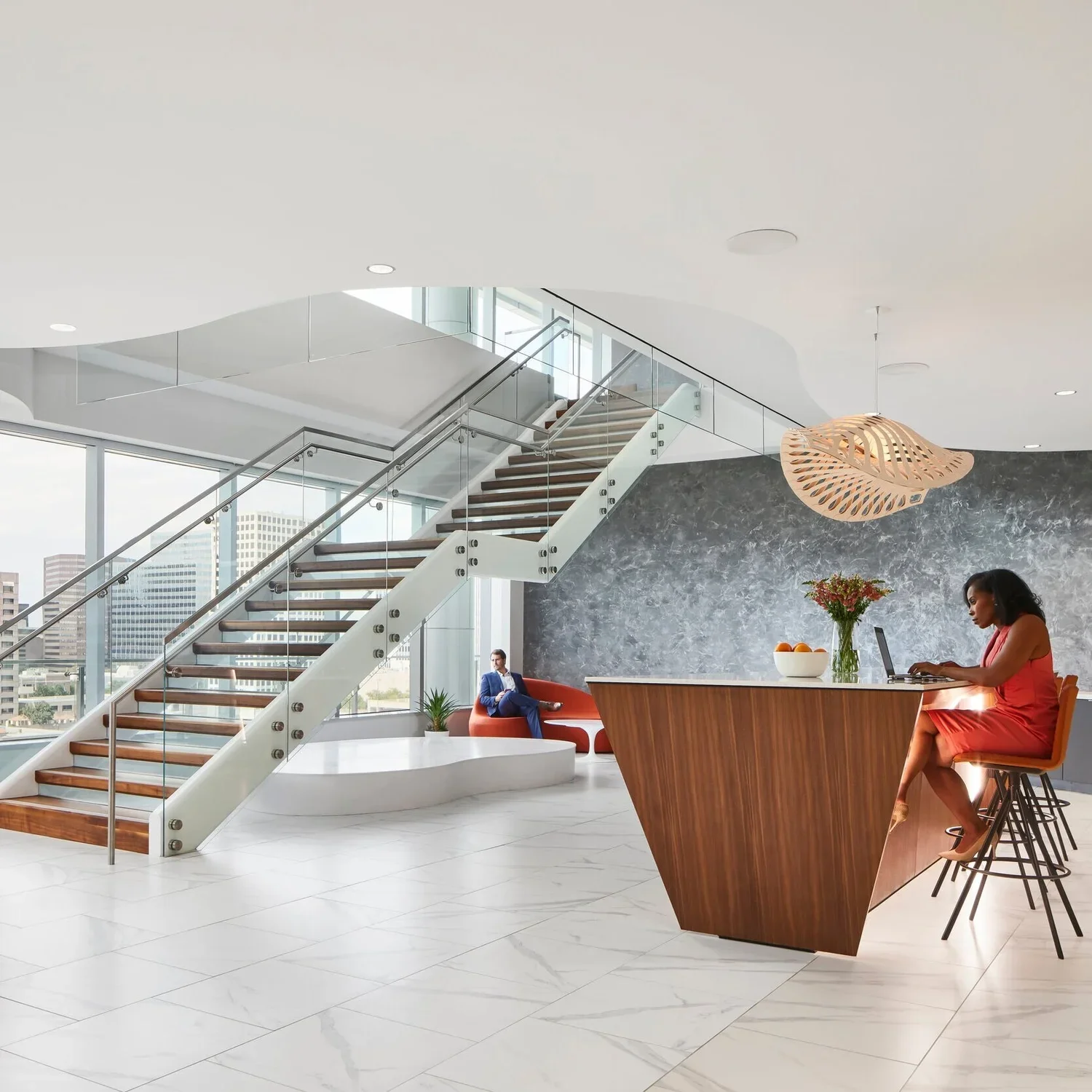 Modern office lobby with a staircase, large windows, and minimalist decor. A woman working on a laptop sits at a wooden counter, while a man sits on an orange chair near the window.