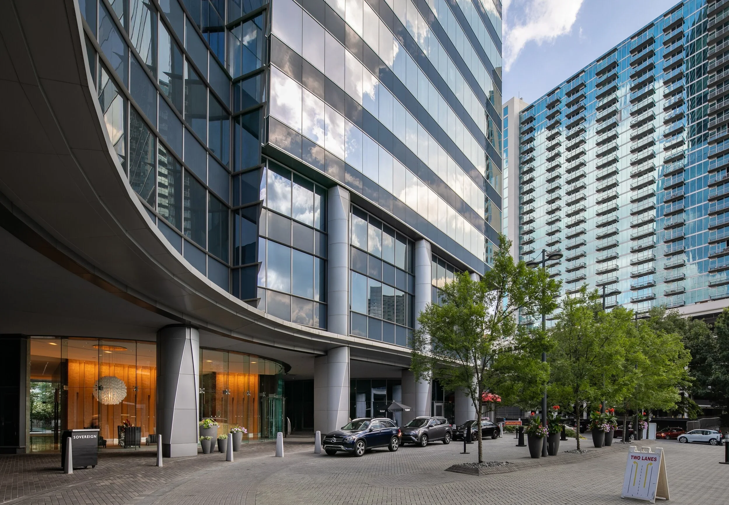 Modern glass office buildings with reflections of the sky and clouds, trees, parked cars, and a street-level entrance with lit interior and potted plants outside.