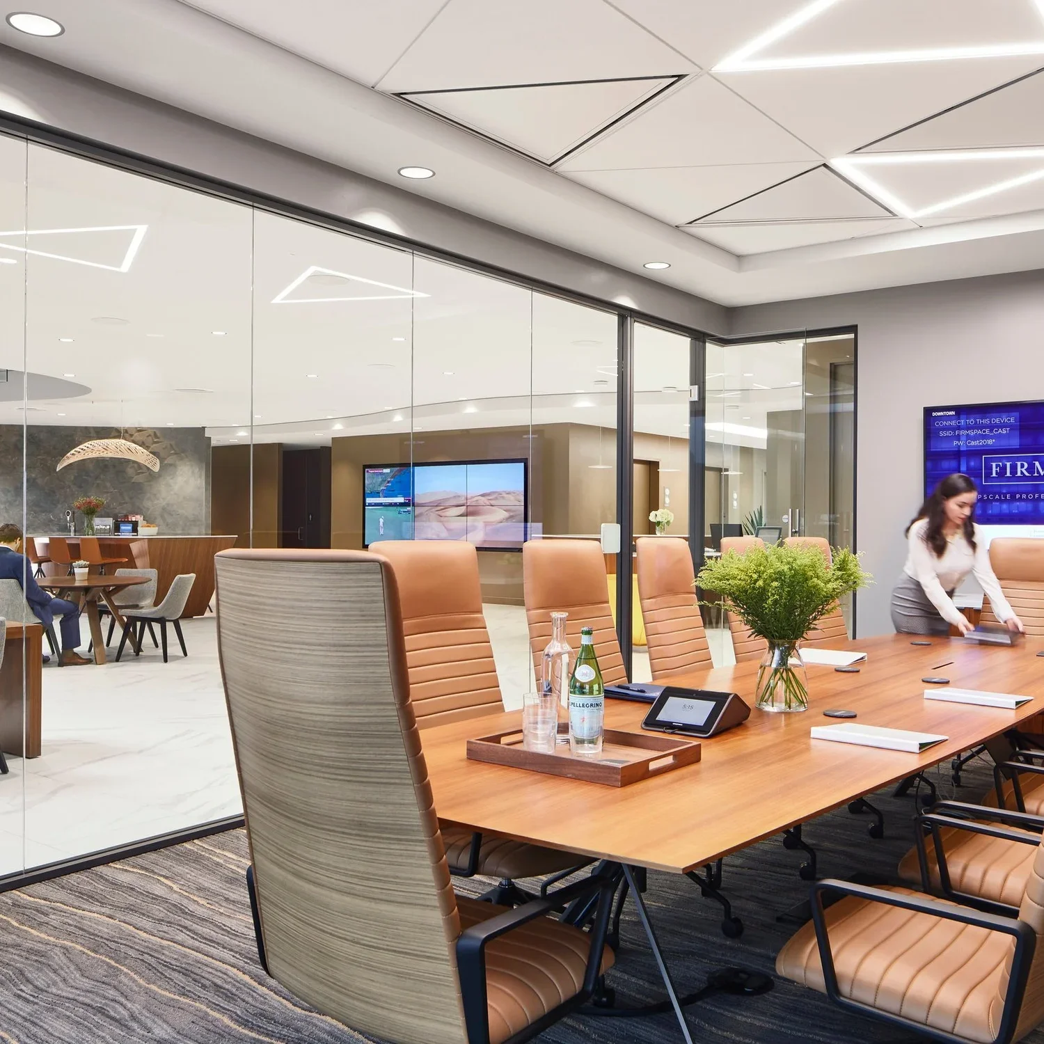 A modern conference room with a large wooden table surrounded by tan leather chairs. There are water bottles, a plant, and a digital conference phone on the table. In the background, a woman is arranging papers, and there are glass walls showing a lo
