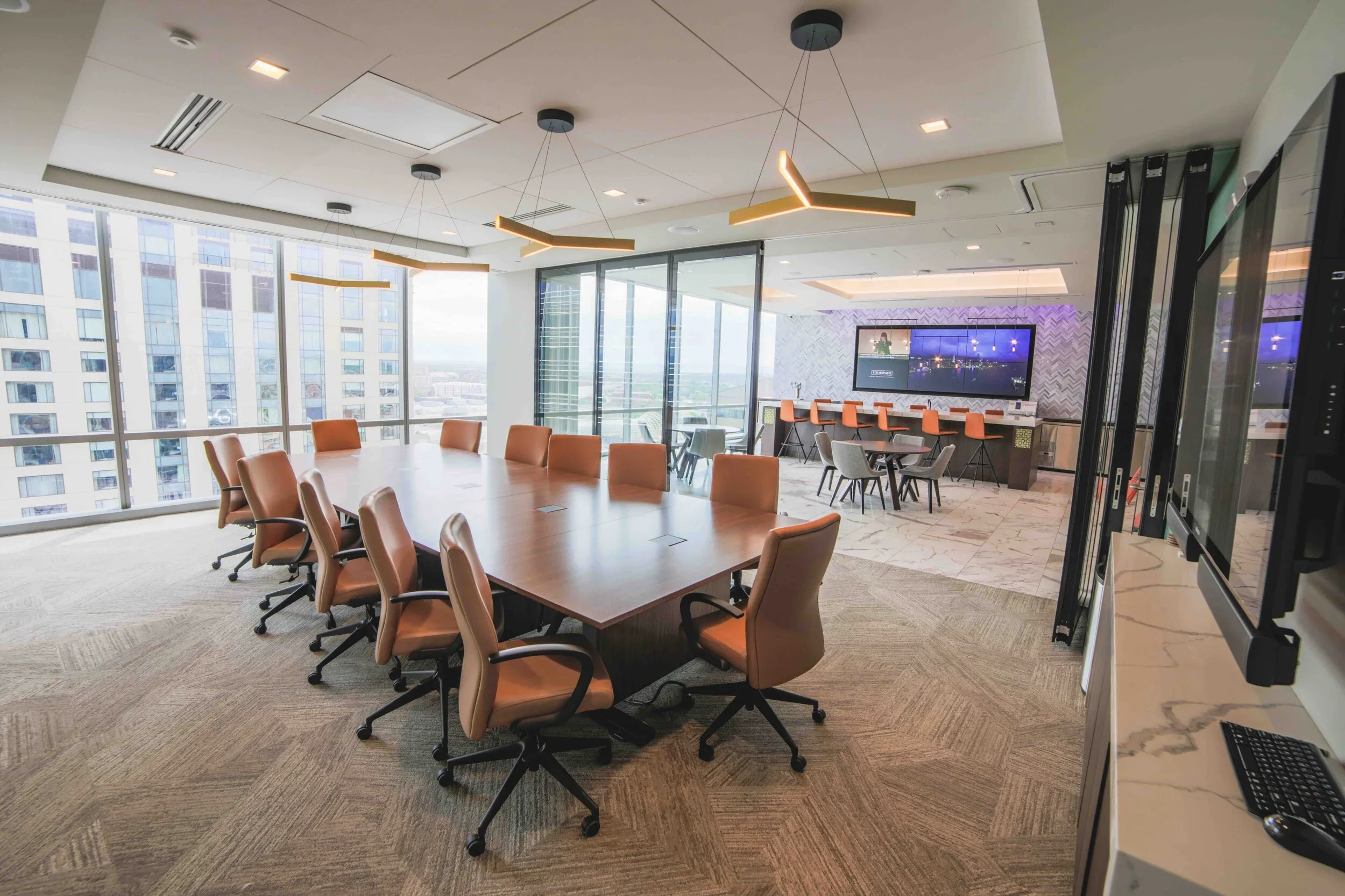 Modern conference room with a large oval table surrounded by tan office chairs, large windows with city view, a bar height counter with orange chairs, and large wall-mounted screens.