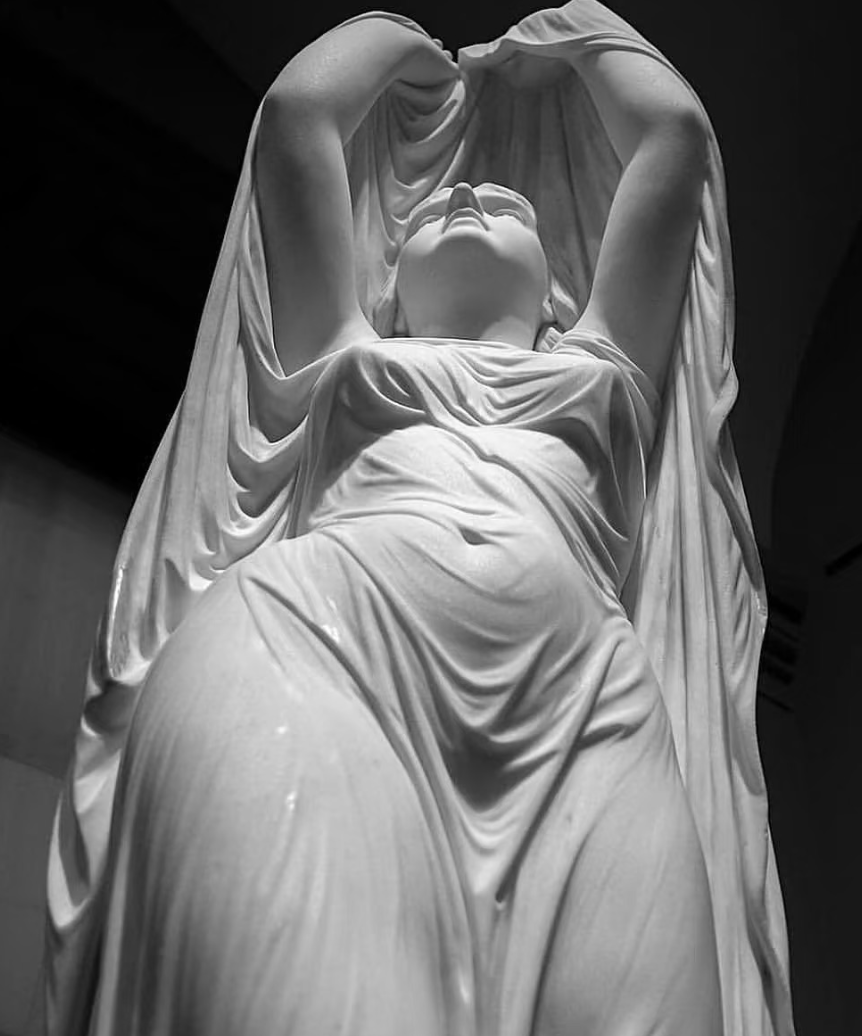 A black and white photograph of a large classical marble sculpture of a woman with her arms raised above her head, draped in flowing garments.