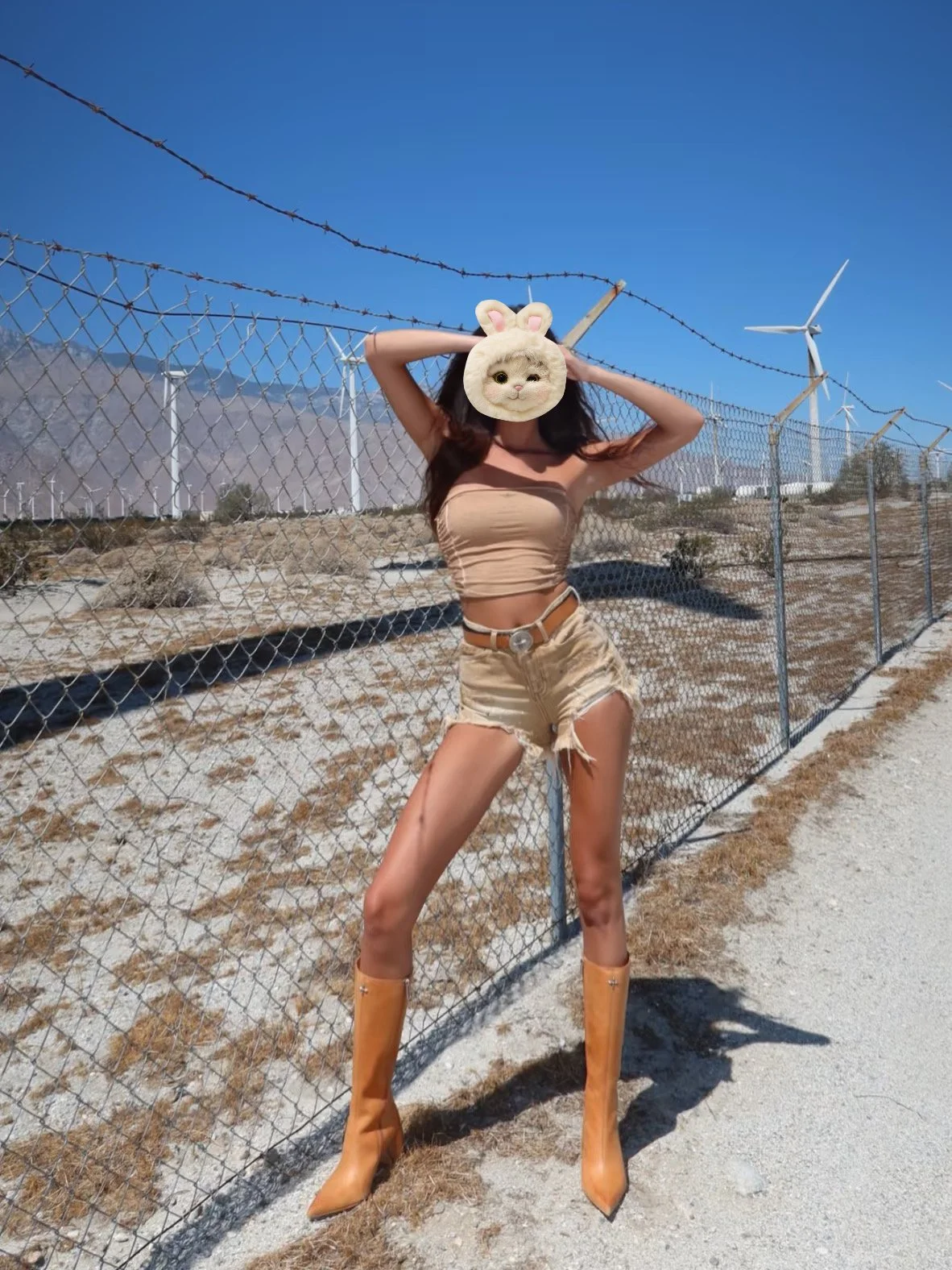 Person standing outdoors on a dirt path near a fence with wind turbines in the background. The person is wearing tan shorts, a tan tube top, tan cowboy boots, and has a cat face mask with a bunny hat graphic over their face.