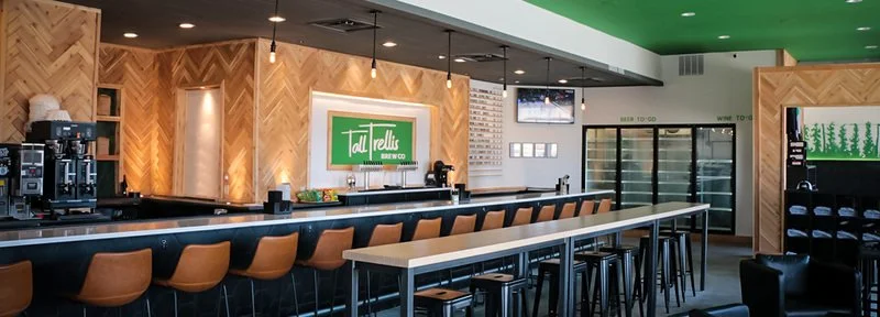 Empty bar area in a restaurant with a sign that says 'Tat Tells Beer Co.' and a row of brown barstools.