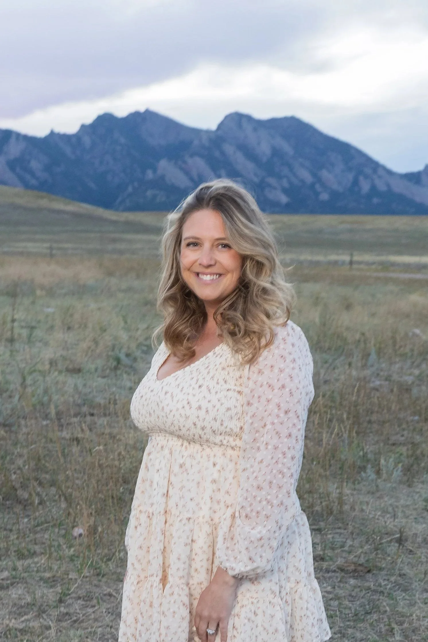 Smiling woman with blonde, wavy hair standing in an open field with mountains in the background.
