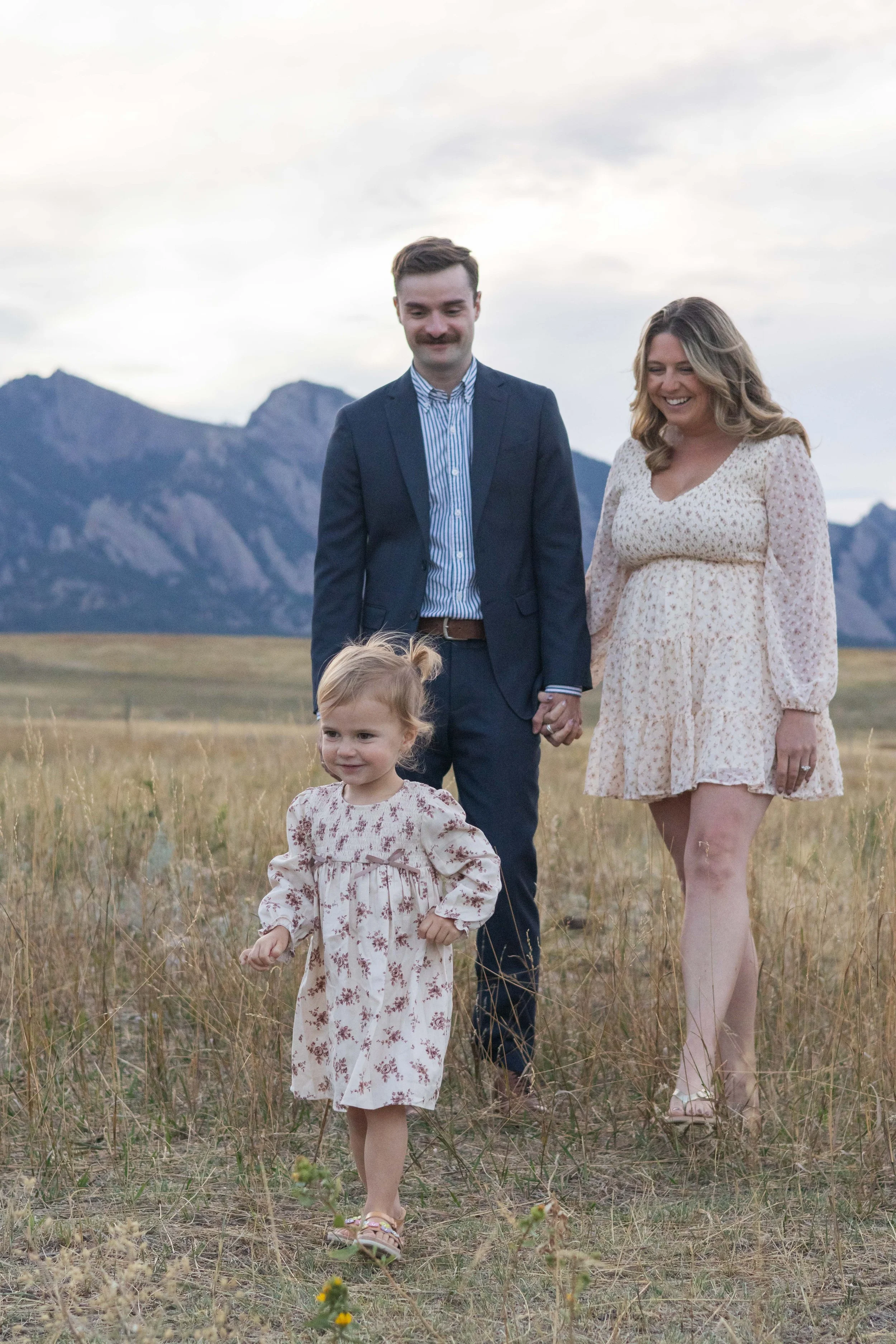 A family of three walking through a grassy field with mountains in the background. The man and woman are holding hands, and a young girl is walking in front of them, all dressed in light, casual clothing.