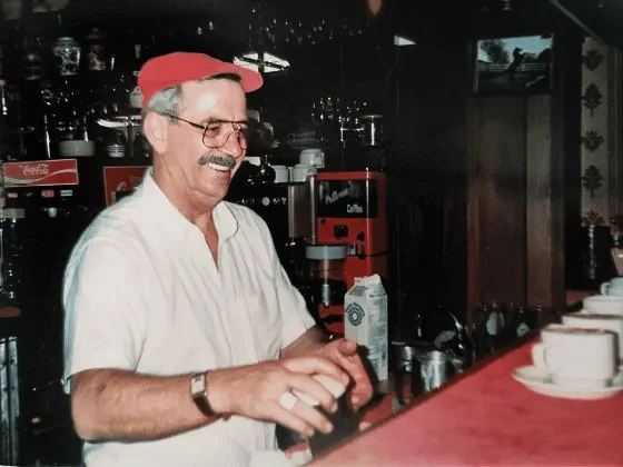 A smiling older man with glasses, gray hair, a red cap, and a white polo shirt, preparing drinks behind a bar counter in a cafe or restaurant.