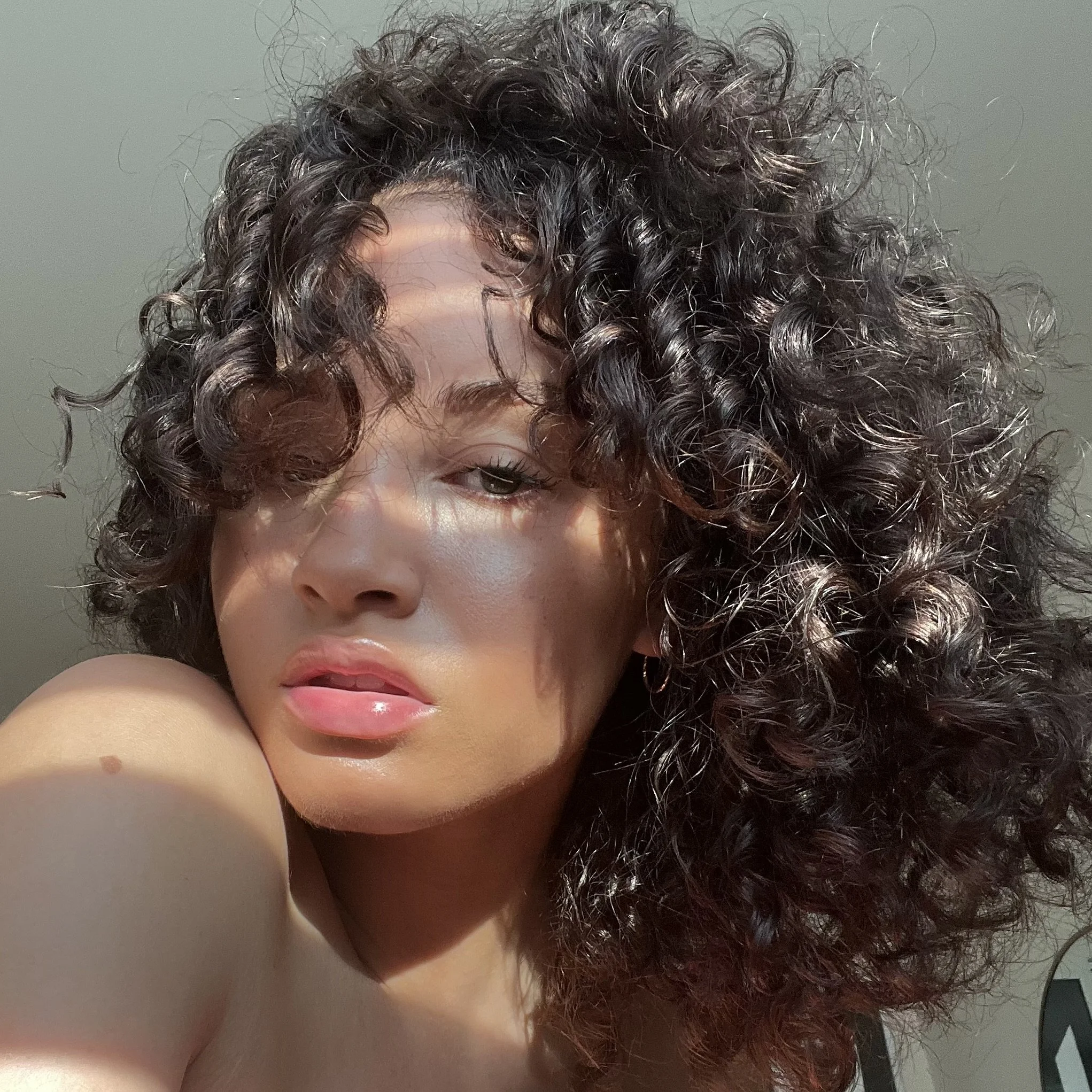 Close-up of a woman with curly dark hair and light skin, looking at the camera with a slightly open mouth, illuminated by sunlight.