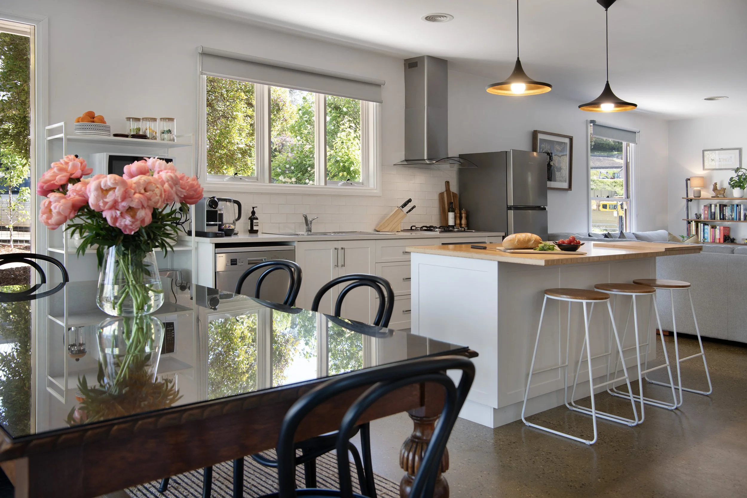 Bright, open-plan kitchen and dining area at Wayward Cottage, featuring a timber island bench, black bentwood chairs, fresh peonies, and garden views through white-framed windows.