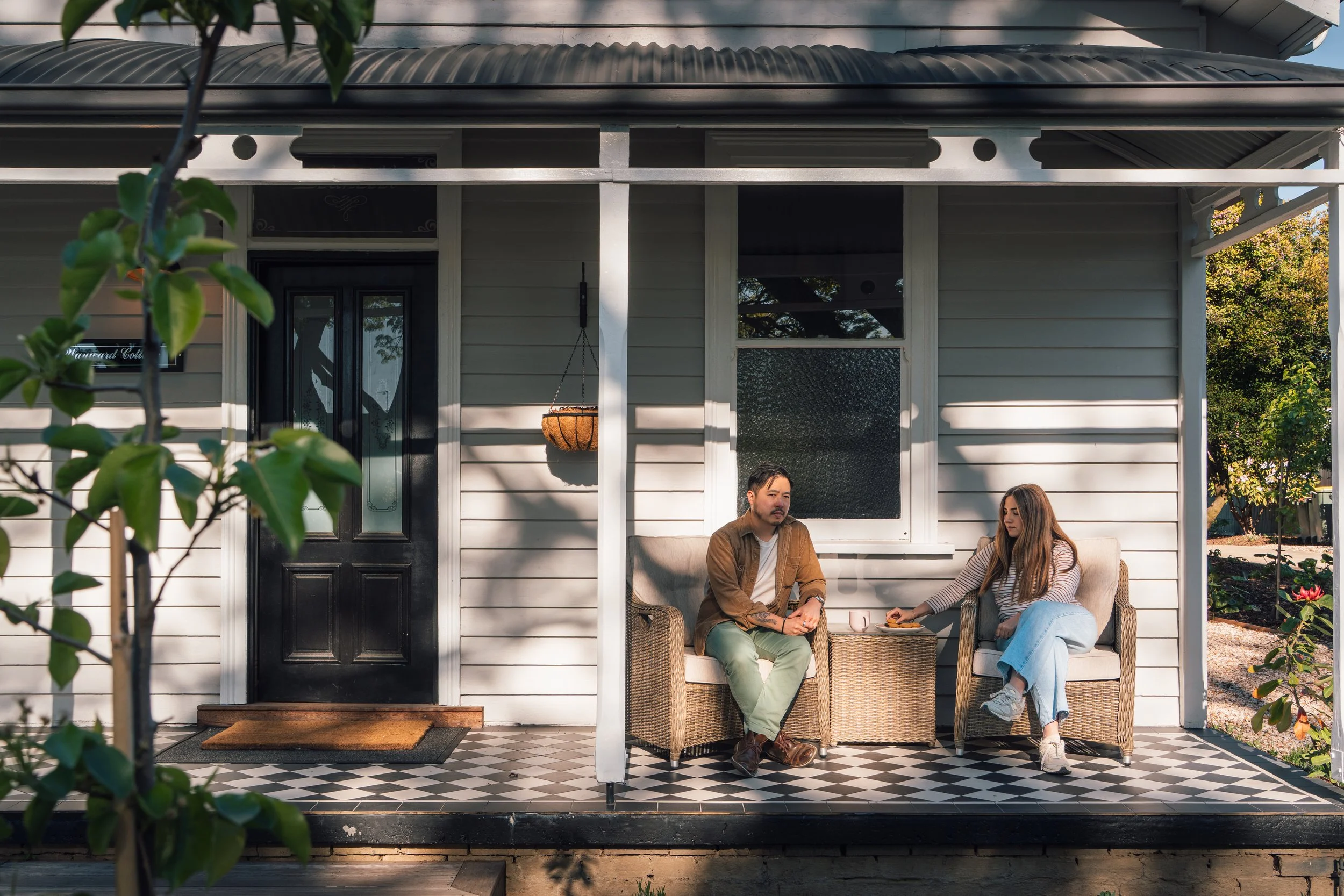 Front porch of a house with two wicker armchairs, a small wicker table, and a checkered black and white tile floor. The house has white siding, a black door, a hanging flower basket, and a small window with a tree visible behind it. There are steps leading up to the porch, and a garden with plants and mulch surrounds it.