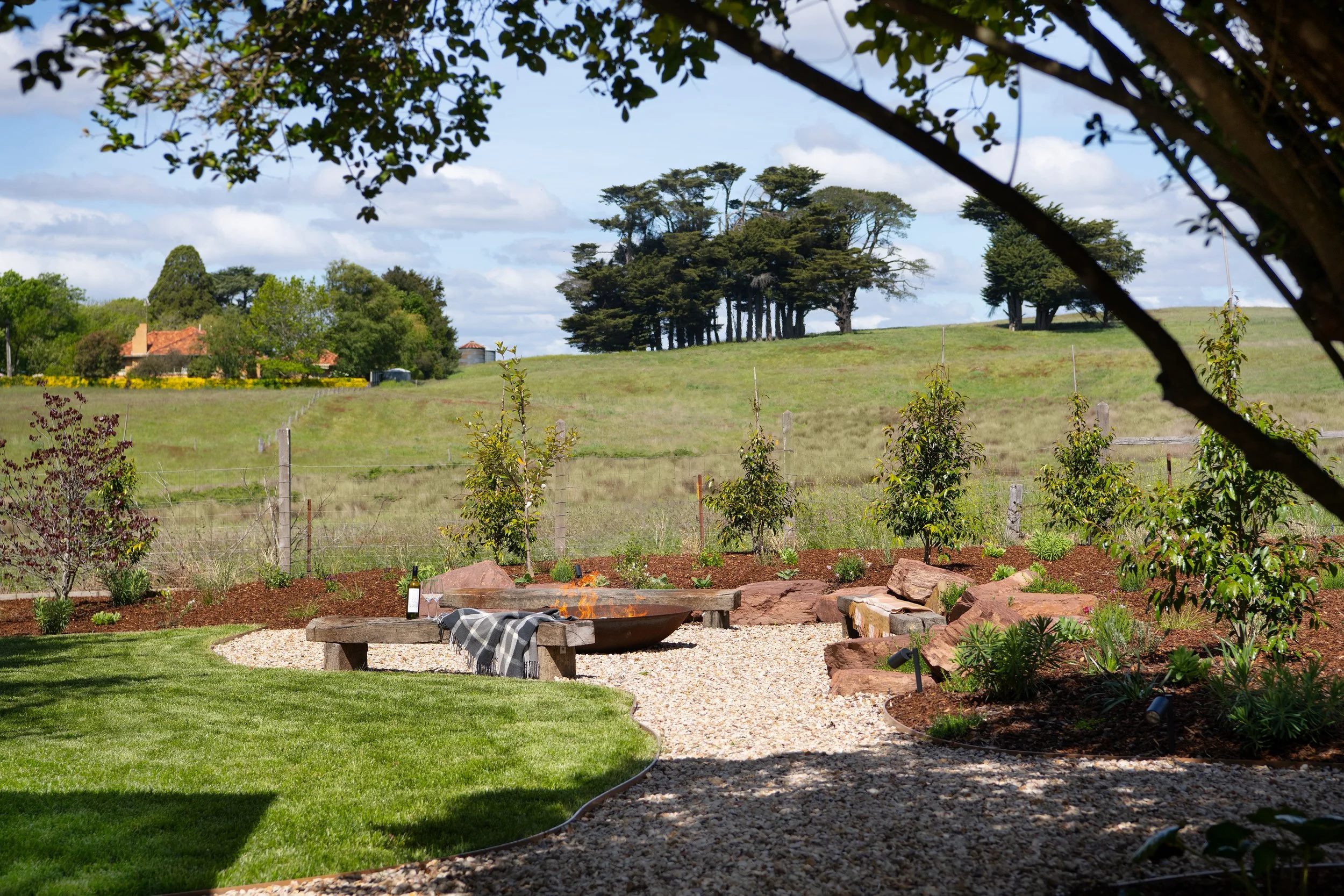A backyard with a fire pit surrounded by two wooden benches on a gravel surface. Small plants and trees are planted along the garden bed, with a grassy area in the foreground. In the background, there is a fence, open fields, and a line of trees under an overcast sky.