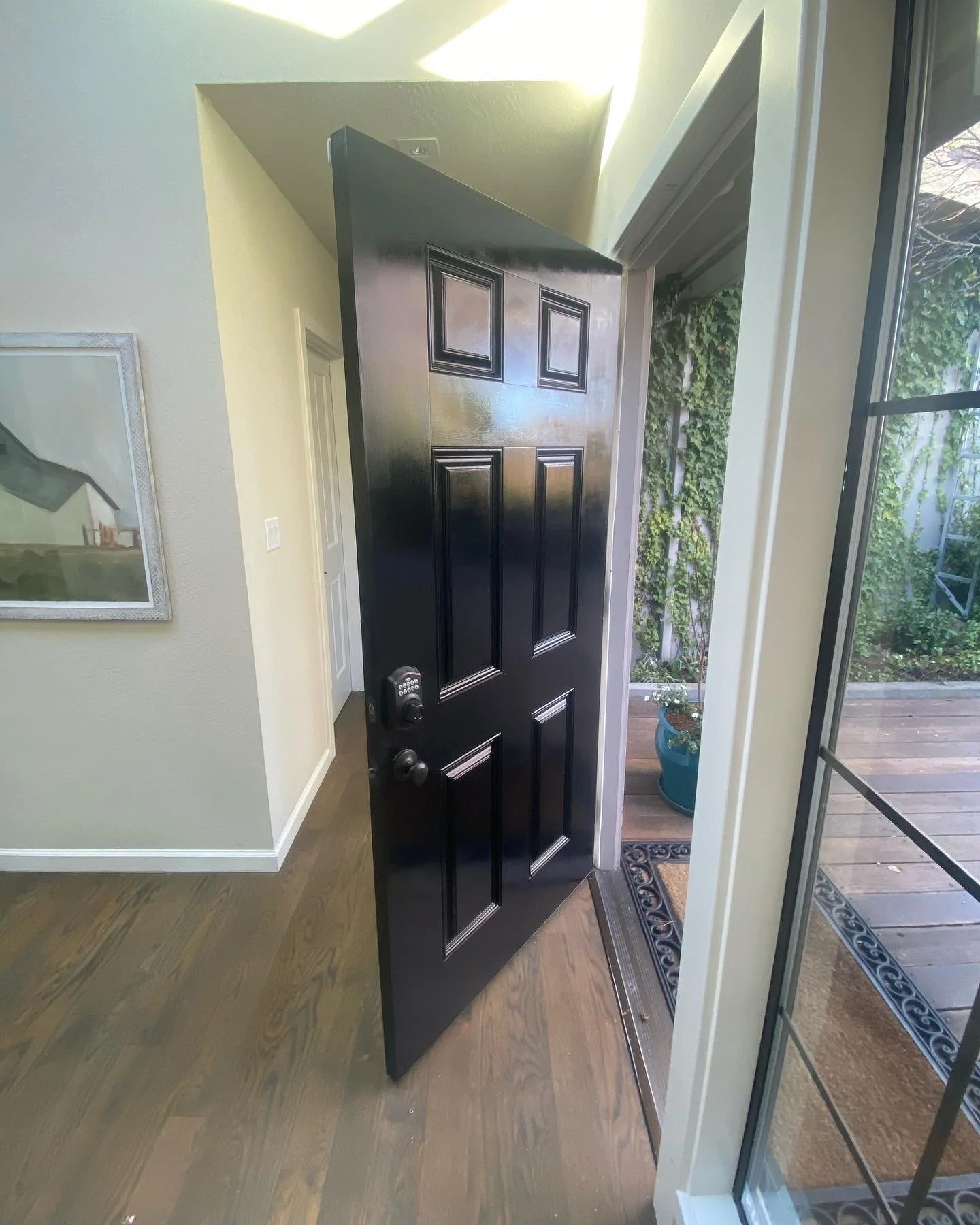 Open black front door with electronic keypad lock, leading to a small porch with wooden decking and a garden outside.