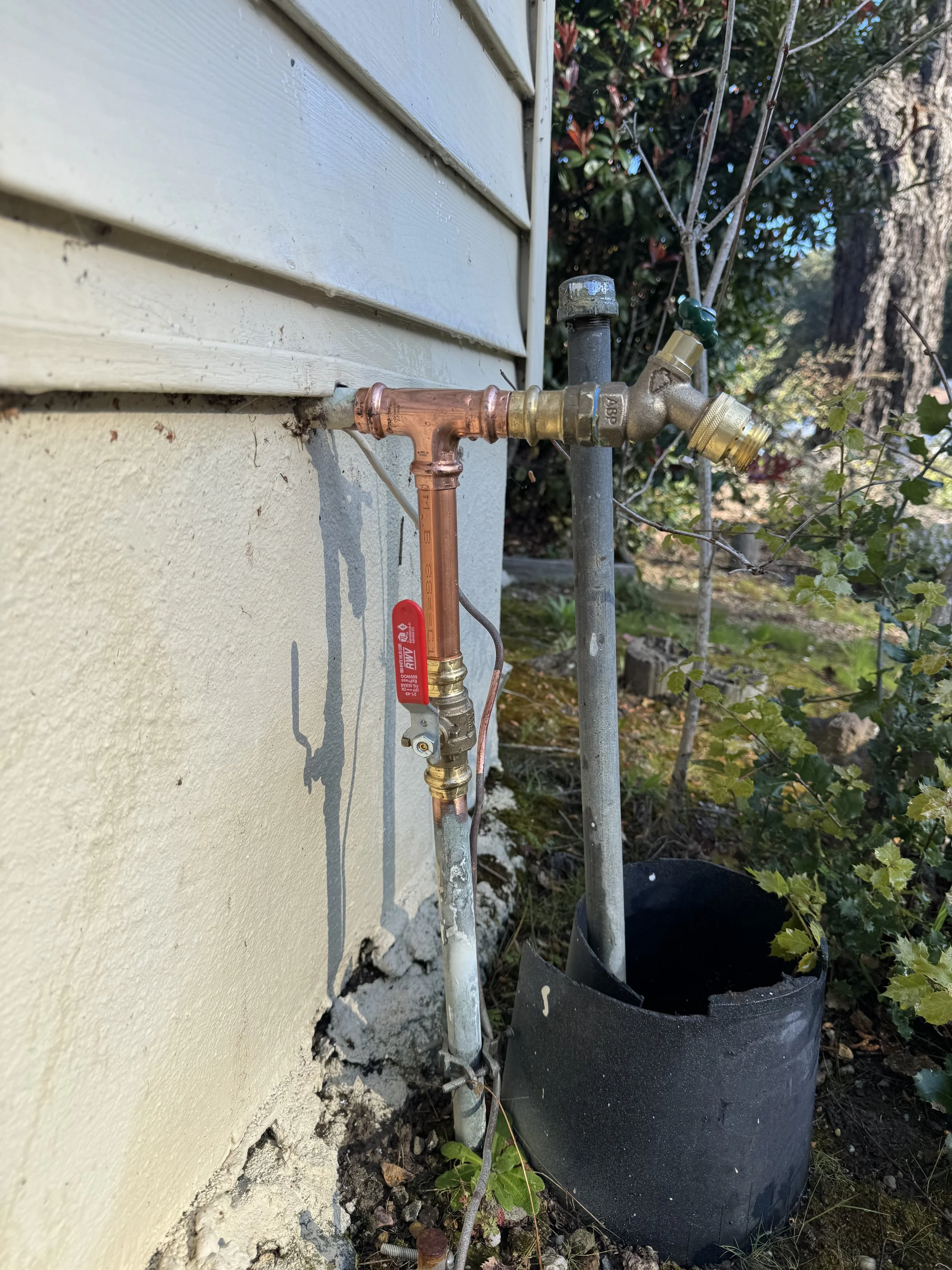 Outdoor water supply setup with copper and brass pipes, a valve, and a black fabric-wrapped pipe covering on a house exterior wall surrounded by garden plants.