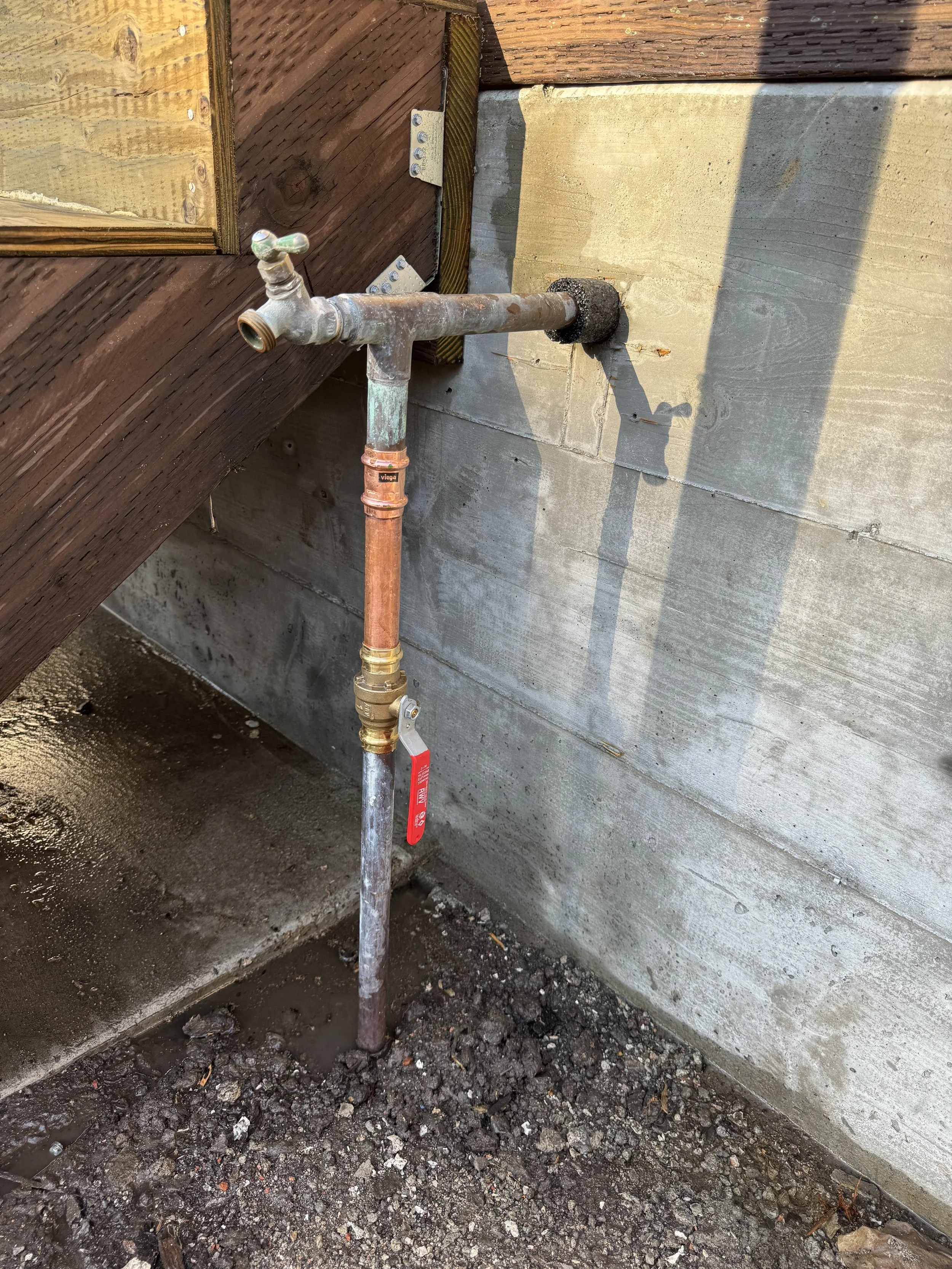 Exterior pipe faucet connected to a copper pipe and partially embedded in concrete, next to a wooden structure and a concrete wall.