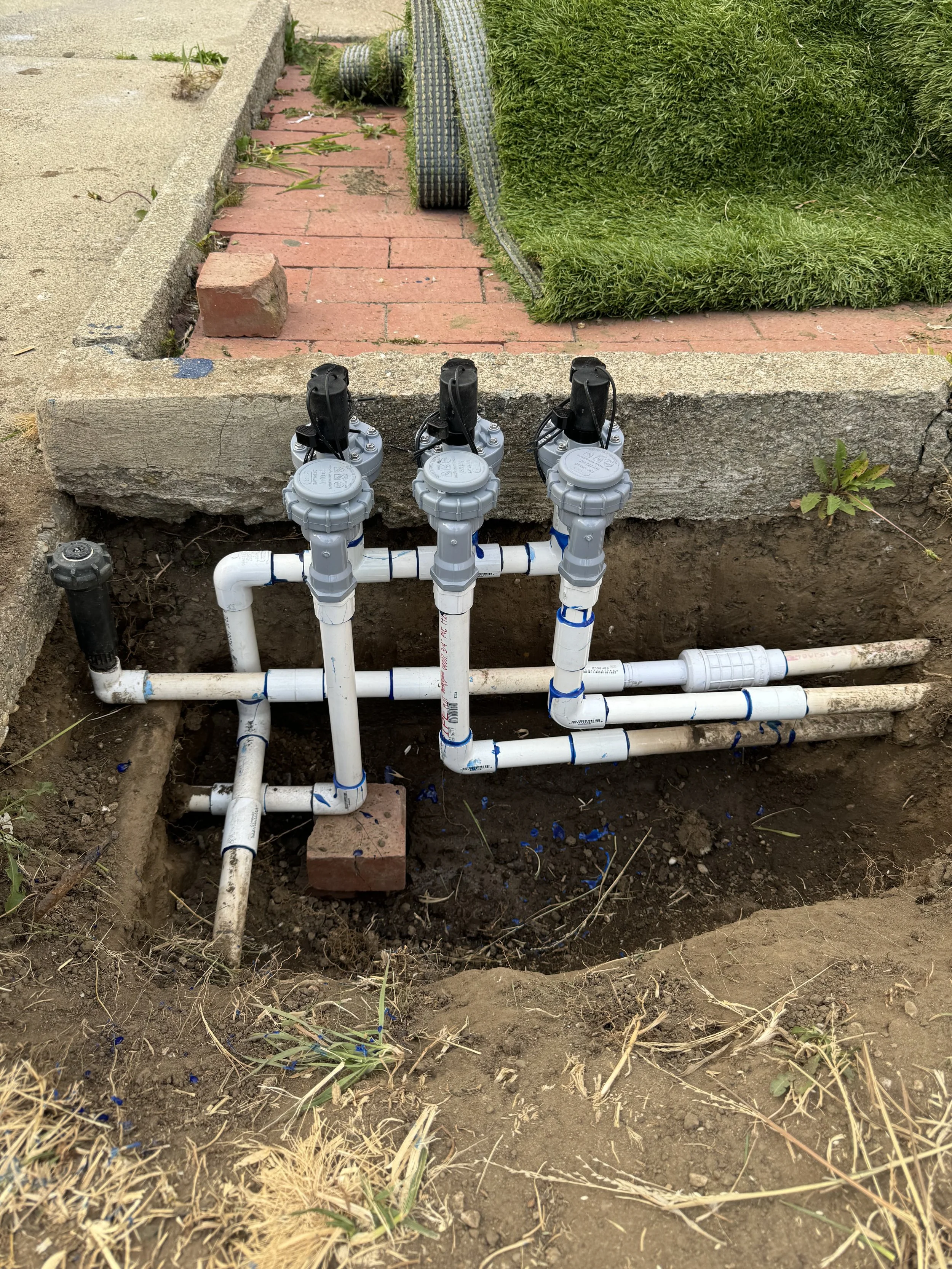 Underground plumbing system with multiple white pipes, valves, and fittings, installed in a dug-out hole near a green grassy area with brick and concrete surfaces nearby.