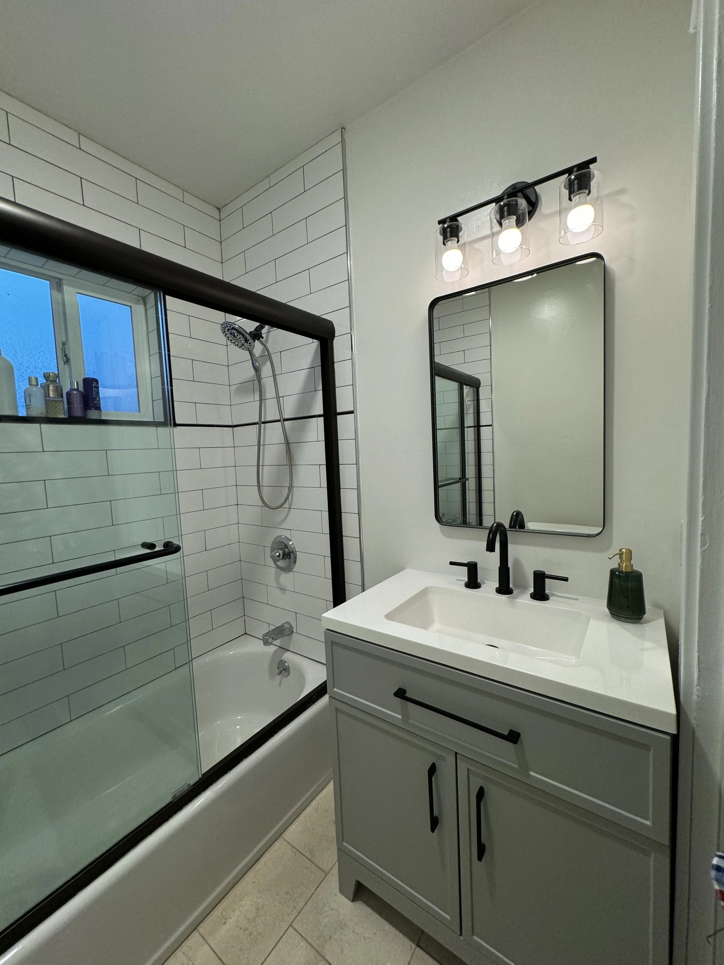 Modern bathroom with white vanity, black faucet, large mirror, light fixture, and a bathtub with sliding glass door with black trim. There are toiletries on the window sill and a small soap dispenser on the sink.