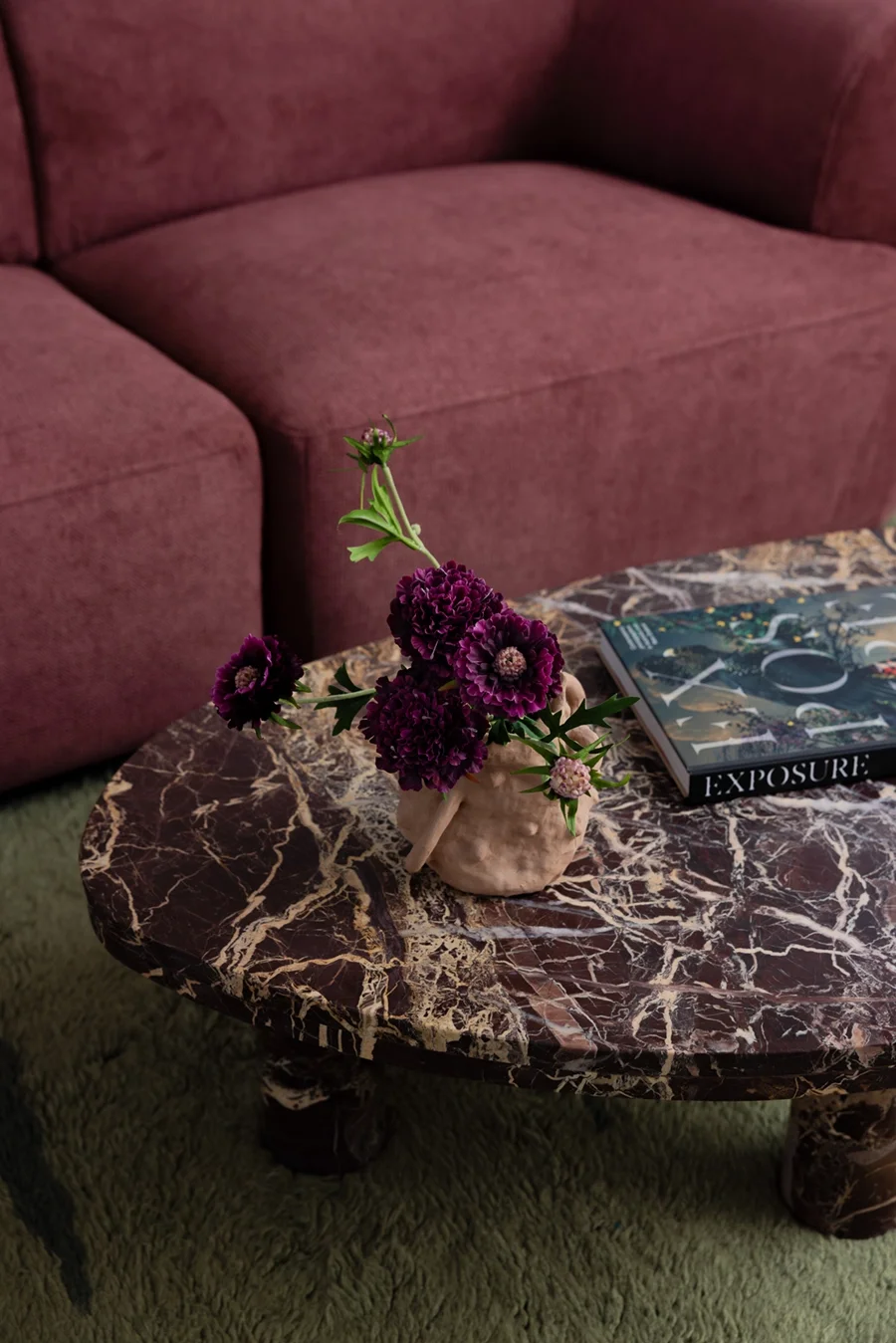 A marble top coffee table with a pink and black pattern, a vase with dark purple flowers, and a magazine titled 'EXPOSURE', placed in front of a pink sofa on a green carpet.