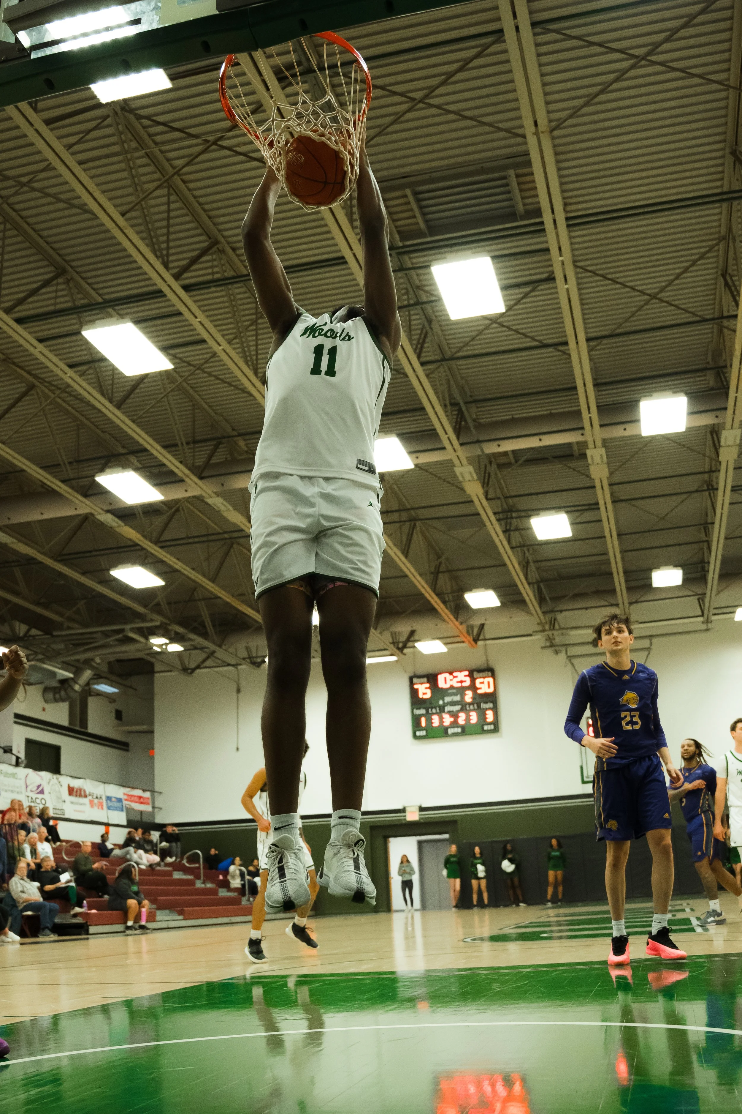 A basketball player in a white jersey with the number 11 is jumping high to dunk the ball into the hoop during a game. Other players and spectators are visible in the background on an indoor basketball court.